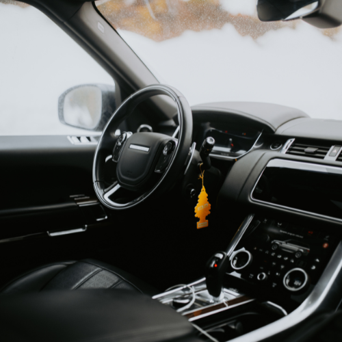 Inside view of a car's dashboard and steering wheel, with a yellow tree-shaped air freshener hanging from the rearview mirror.