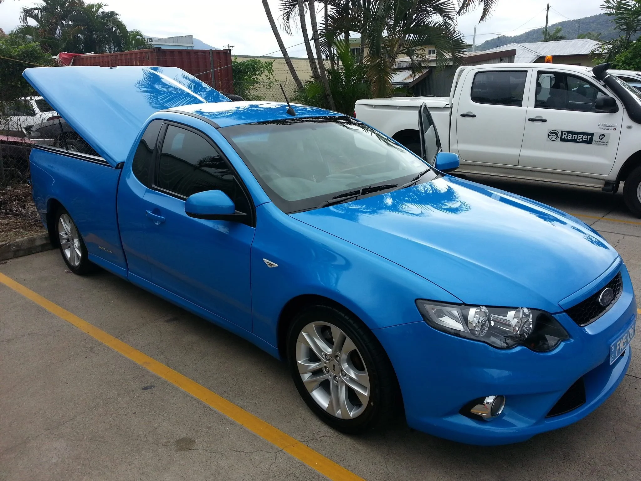 A blue pickup truck with its hood open parked next to a white ranger vehicle in an outdoor parking lot, with palm trees and buildings in the background.