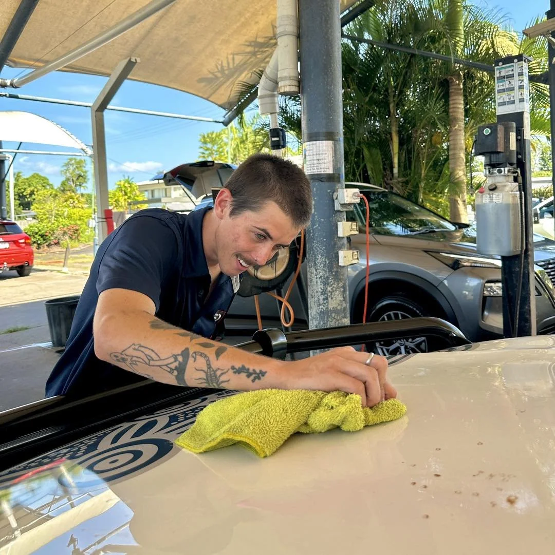 A young man with tattoos on his arm is polishing the hood of a yellow car with a yellow towel at an outdoor car wash station.