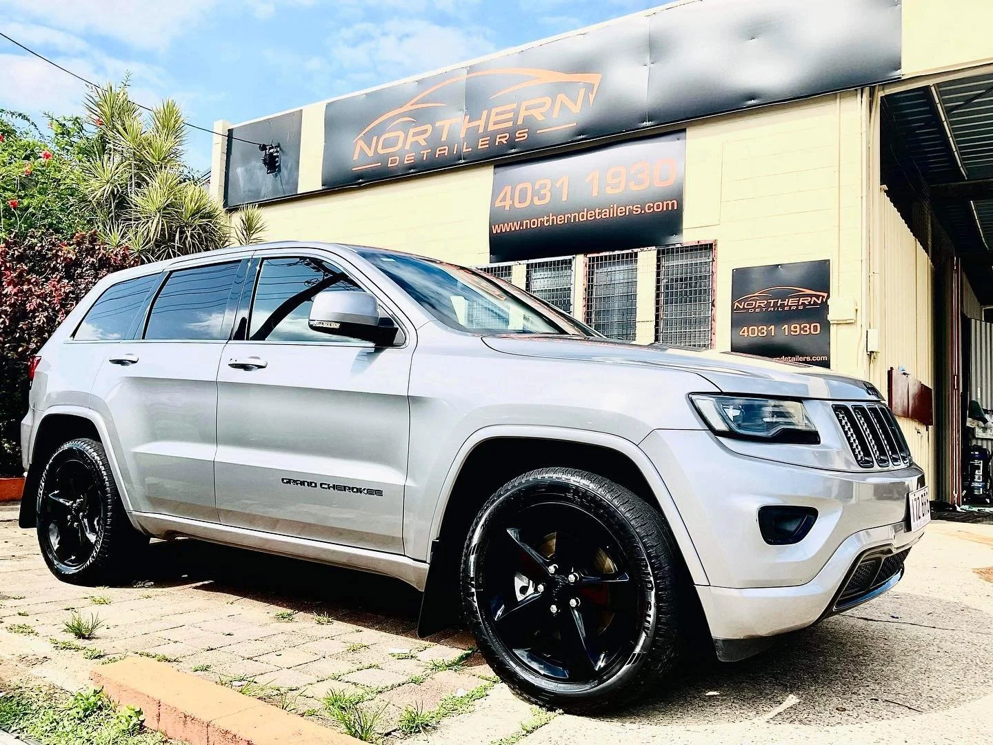 Silver Jeep Grand Cherokee parked in front of Northern Detailing car repair shop with signage, plants, and blue sky in background.