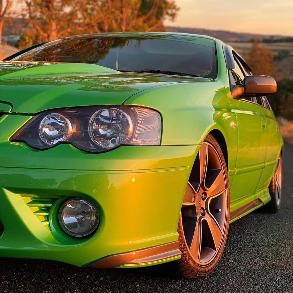A bright green sports sedan parked on the road during sunset with trees in the background. The car has large alloy wheels and sleek design.
