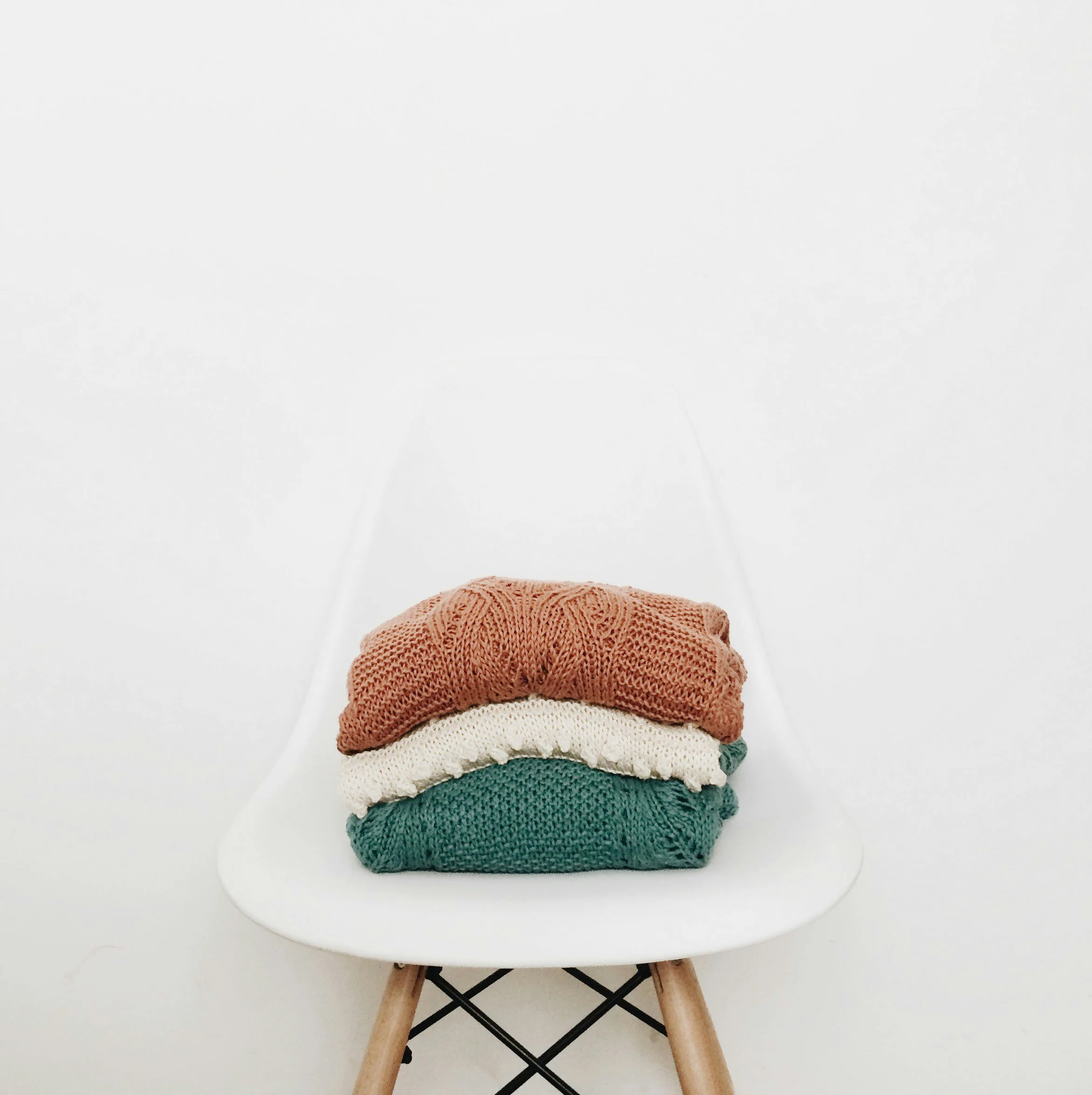 Three folded knitted sweaters in brown, white, and green placed on a modern white chair with wooden legs against a plain white background.