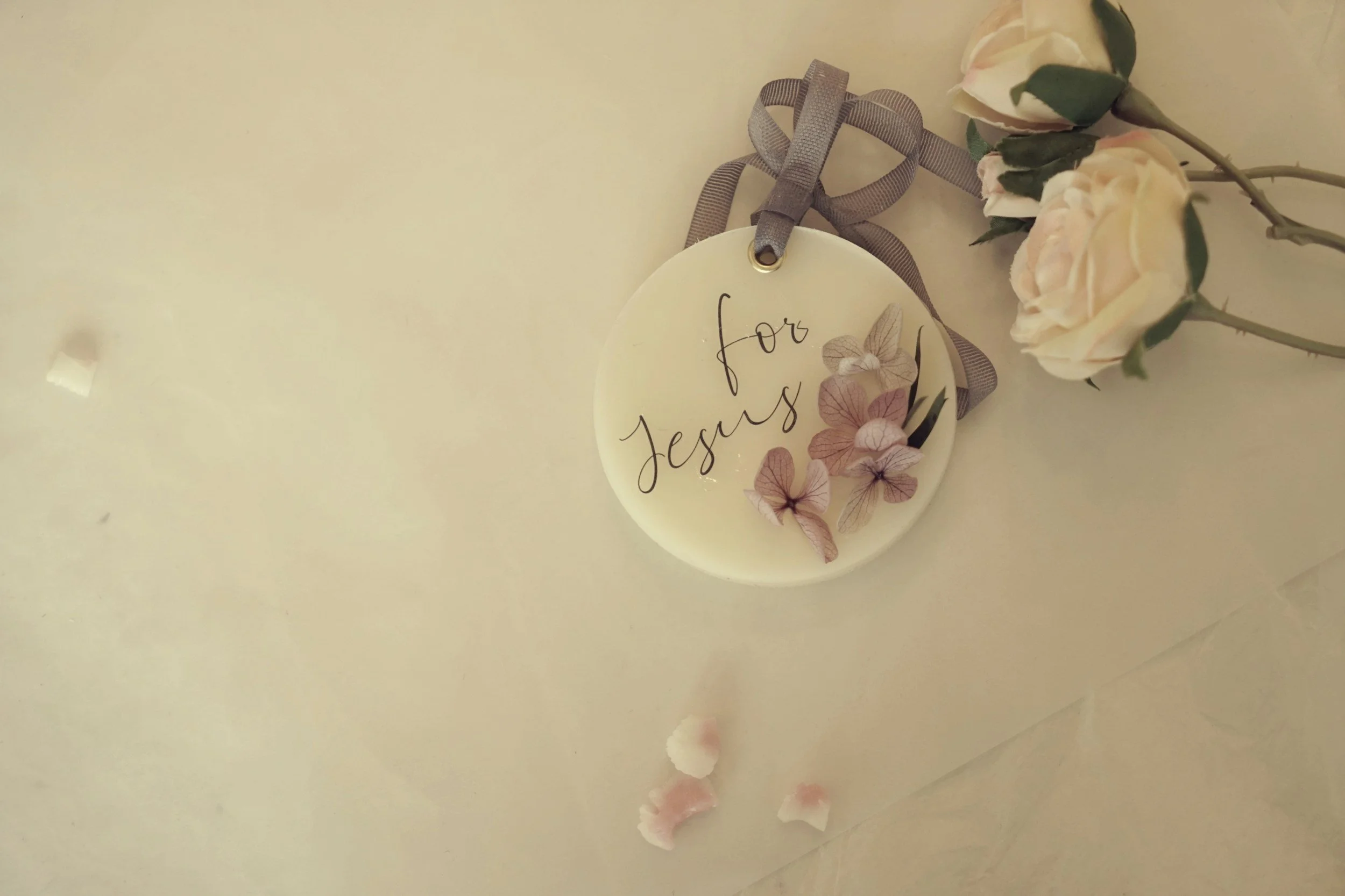 A white decorative plaque with the words 'for Jesus' written in cursive, decorated with pink and beige paper flowers and a gray ribbon, placed on a light surface next to a bouquet of white roses.