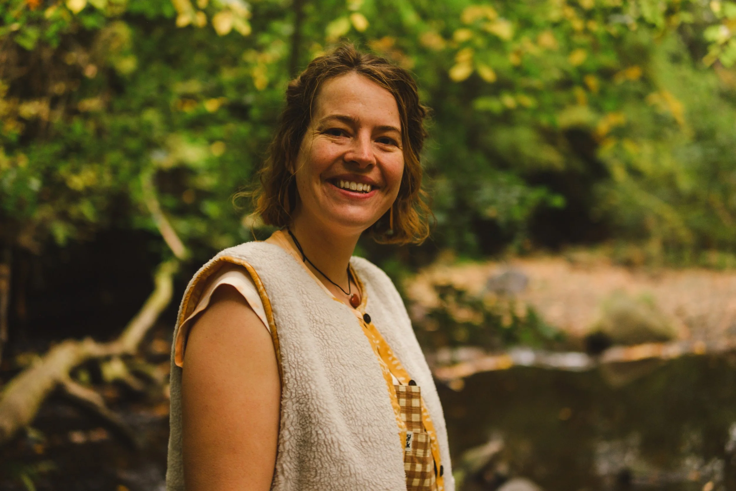 A woman with shoulder-length brown hair smiling outdoors among green trees.