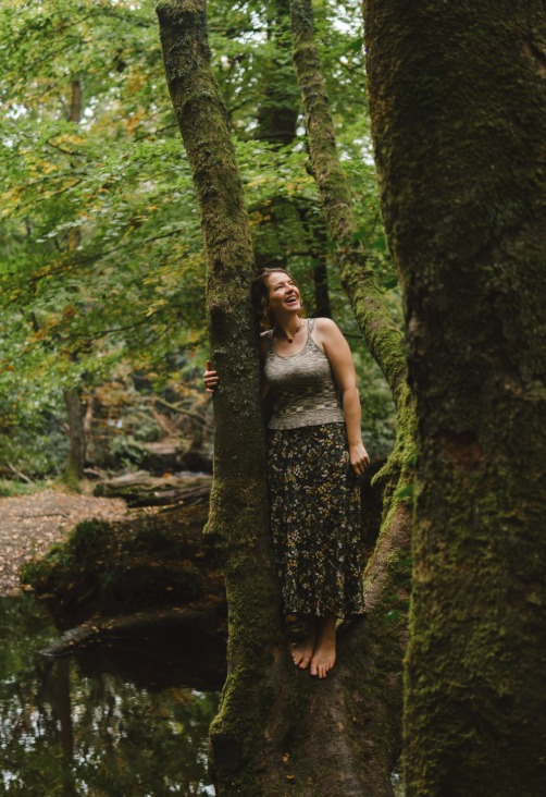 A woman standing on a moss-covered tree trunk in a lush green forest, smiling and looking up amidst trees and foliage.