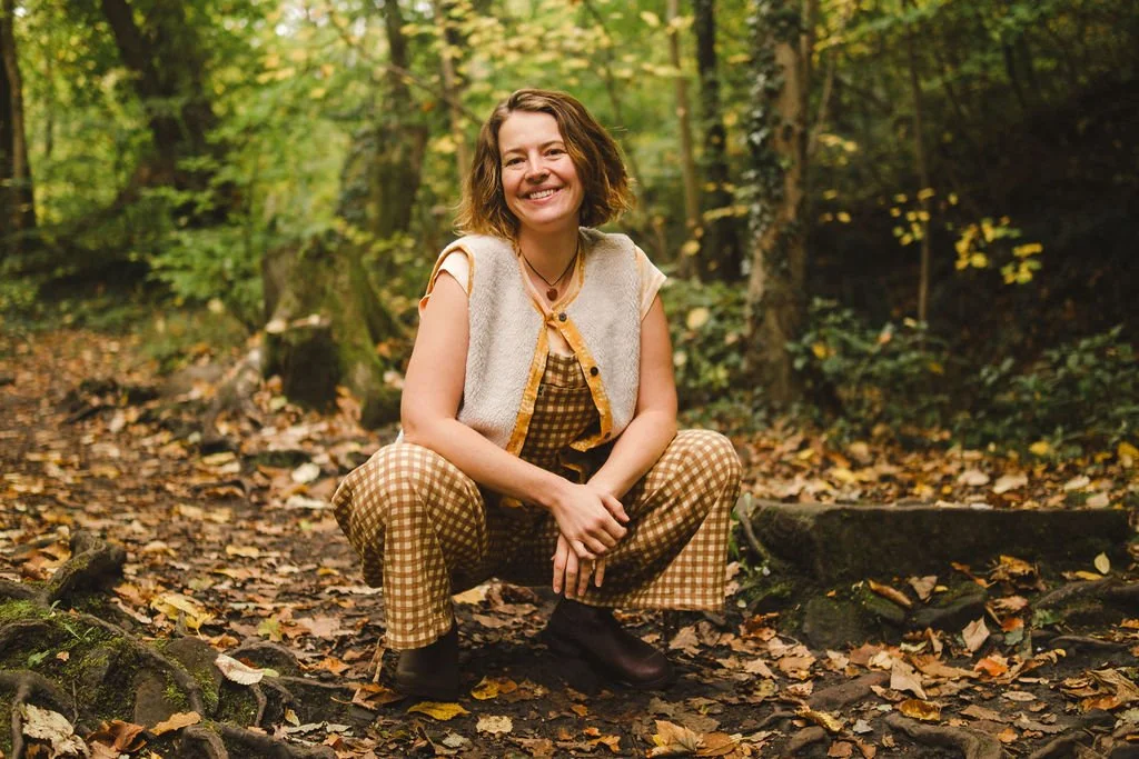 Young woman squatting in a forest with autumn leaves on the ground, smiling, wearing a beige vest, checked pants, and black boots.