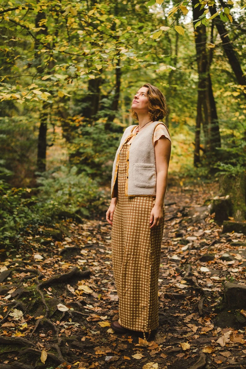 A woman stands on a forest trail surrounded by autumn foliage, looking upward with a peaceful expression.