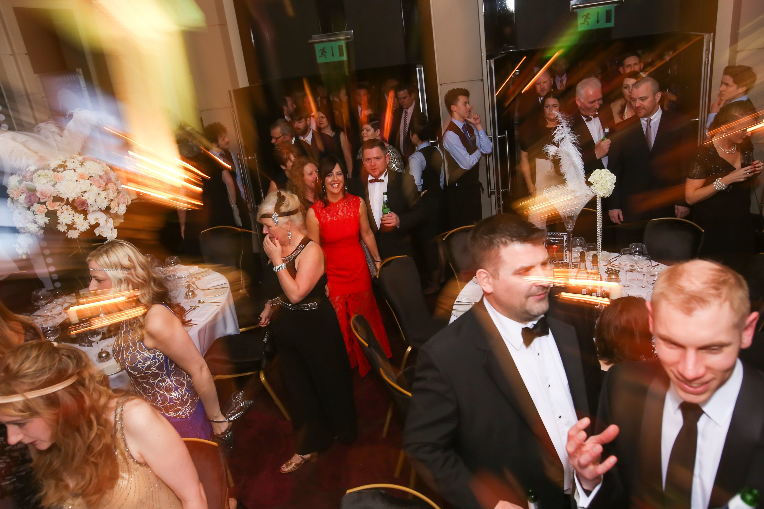 People dressed in formal attire at a lively indoor event, with decorations and lights creating a festive atmosphere.