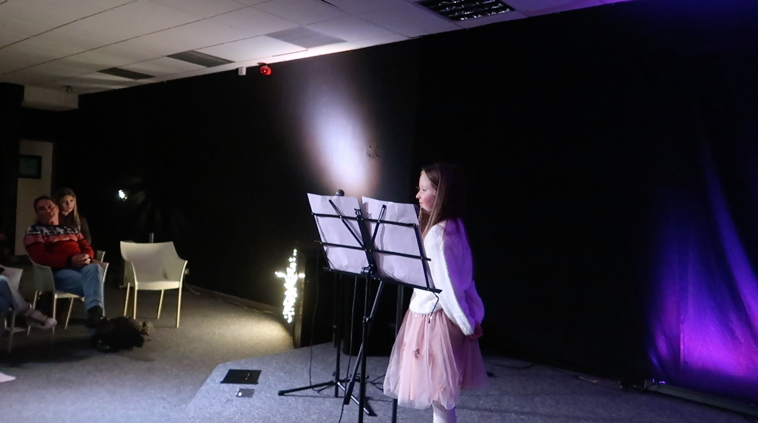 A young girl standing in front of a microphone and music stand, performing on stage with other audience members watching.