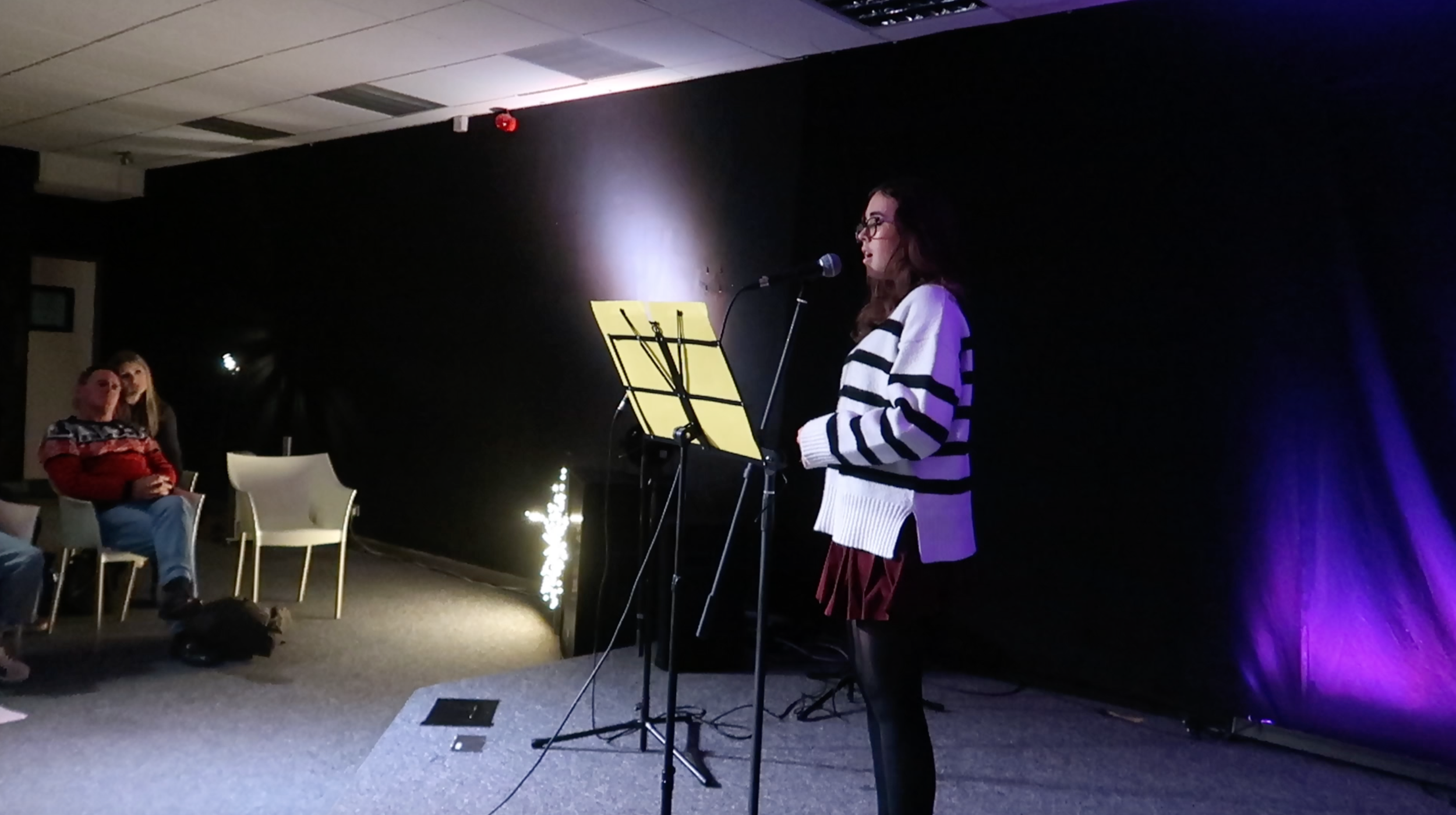 A woman with glasses and long brown hair wearing a white and black striped sweater, red skirt, and black tights stands on stage with a microphone, singing or speaking, in a dark room with a black background and purple lighting.