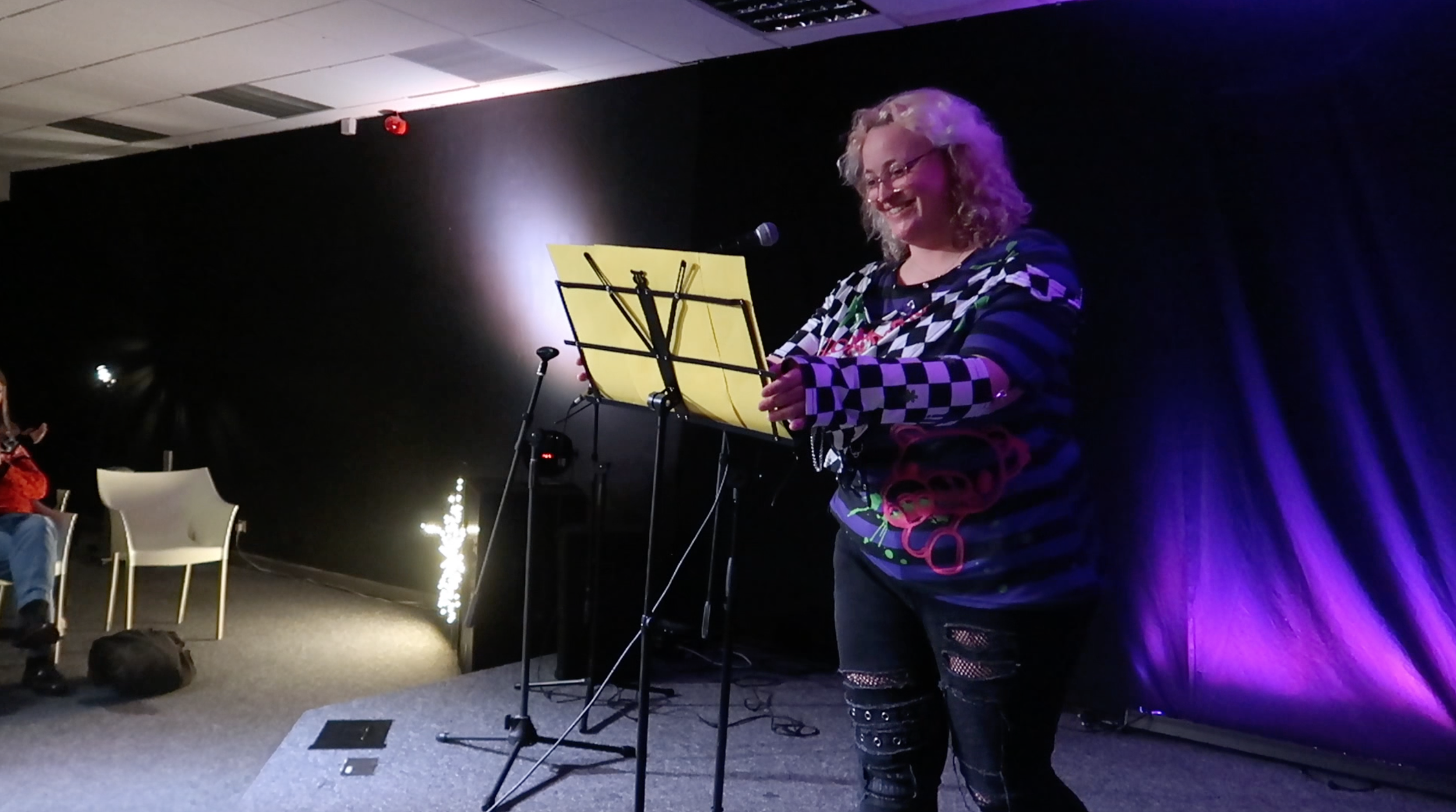 A woman with curly blonde hair and glasses standing on stage, smiling, reading from a yellow music stand, with a microphone in front of her and stage purple lighting in the background.