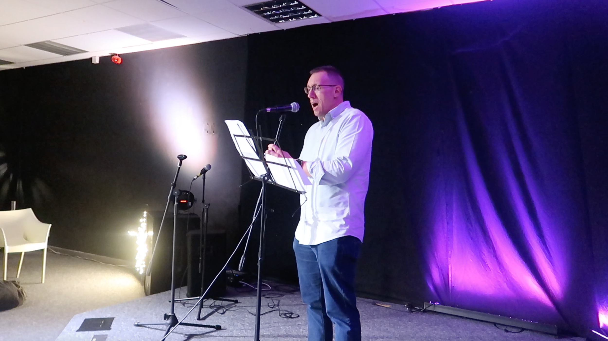 A man in a white shirt and glasses stands on a stage, singing or speaking into a microphone with a music stand in front of him. The stage background is black with purple lighting.