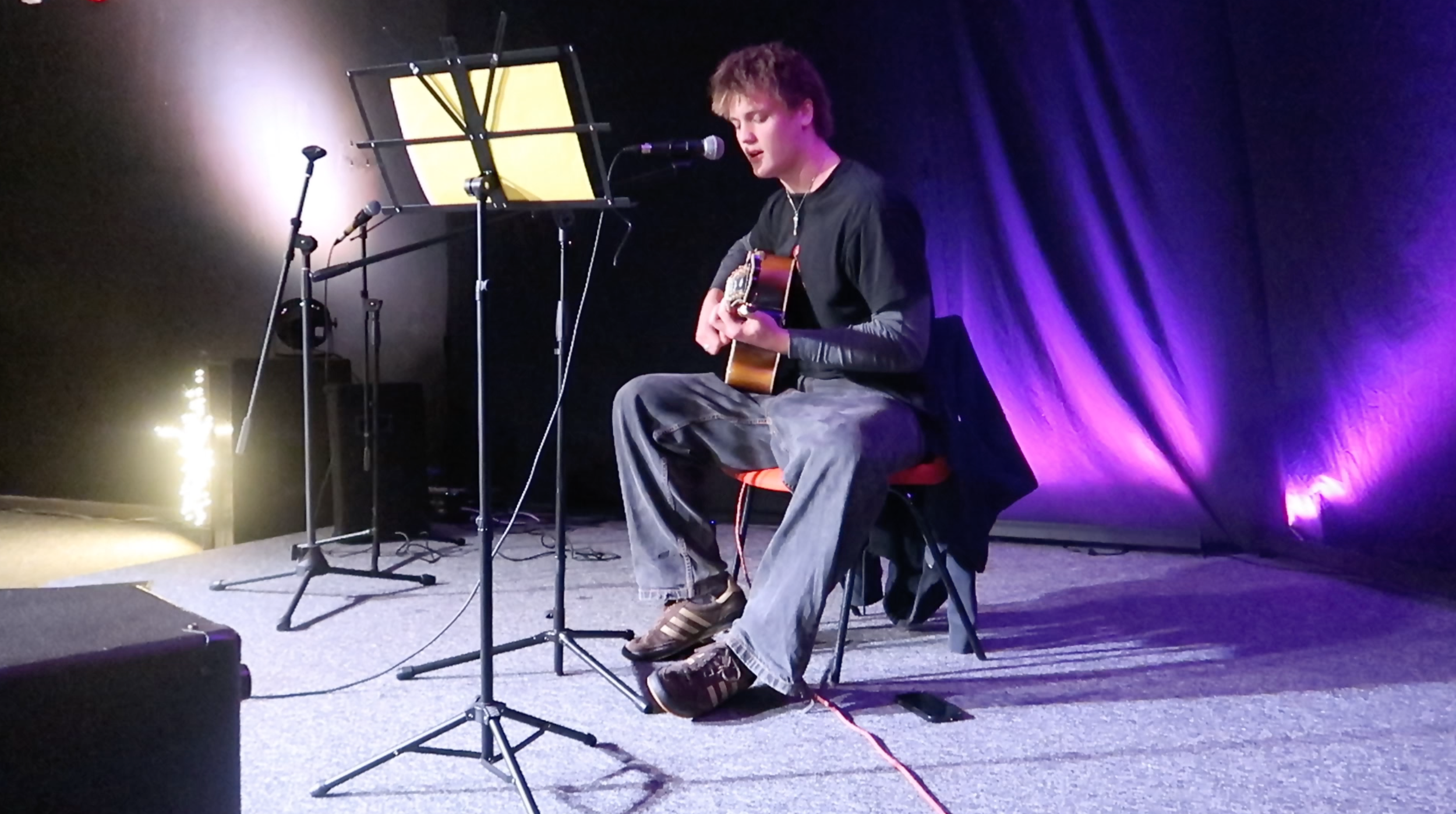 Young man sitting on a stool, playing an acoustic guitar on stage with microphones and music stands around him, purple stage lighting highlighting the scene.