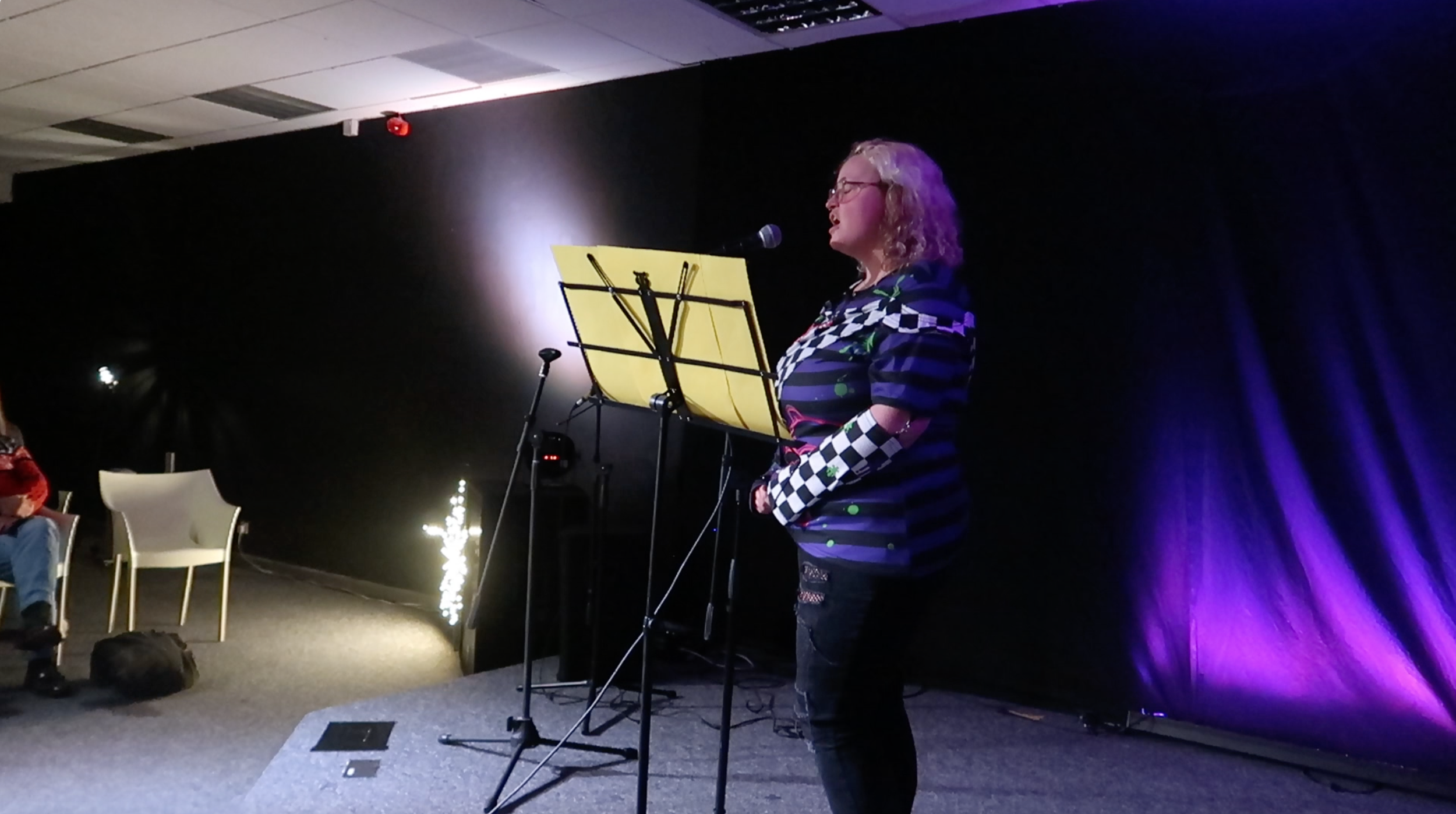 A woman with blonde curly hair and glasses stands on a stage, singing into a microphone with a yellow music stand in front of her. The stage has a black backdrop with purple lighting, and part of the audience with empty chairs is visible on the left.