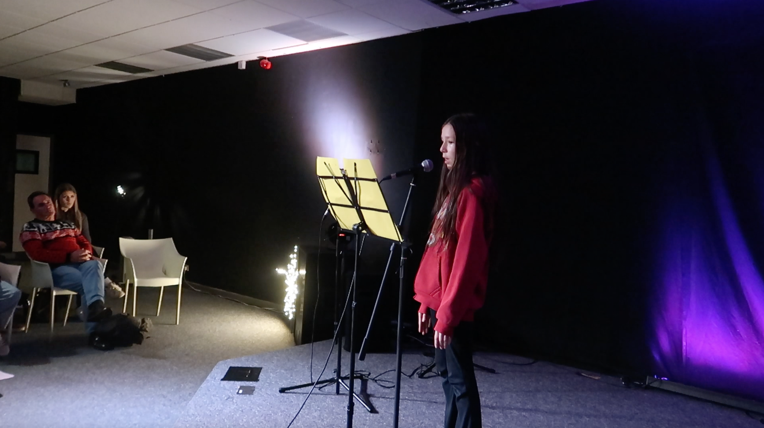 A young woman in a red jacket standing in front of a microphone on stage, reading or singing from a music stand with yellow sheets, in a dark room with purple lighting. Audience members sitting on chairs in the background watch her perform.