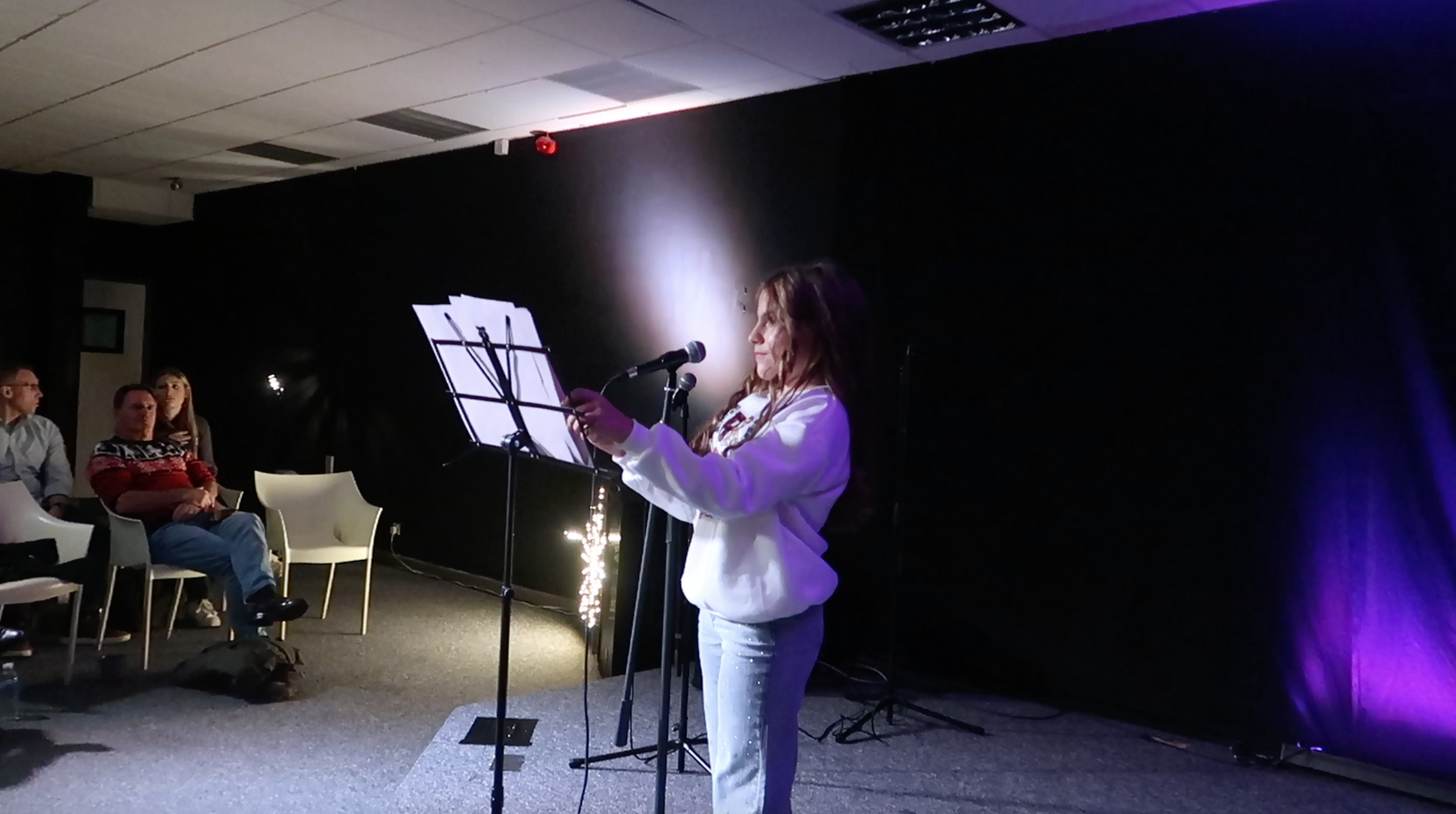 A woman standing on stage, reading from a sheet of paper, with a microphone in front of her. The stage is lit with purple lighting. Audience members are seated on white chairs to the left, watching her perform.