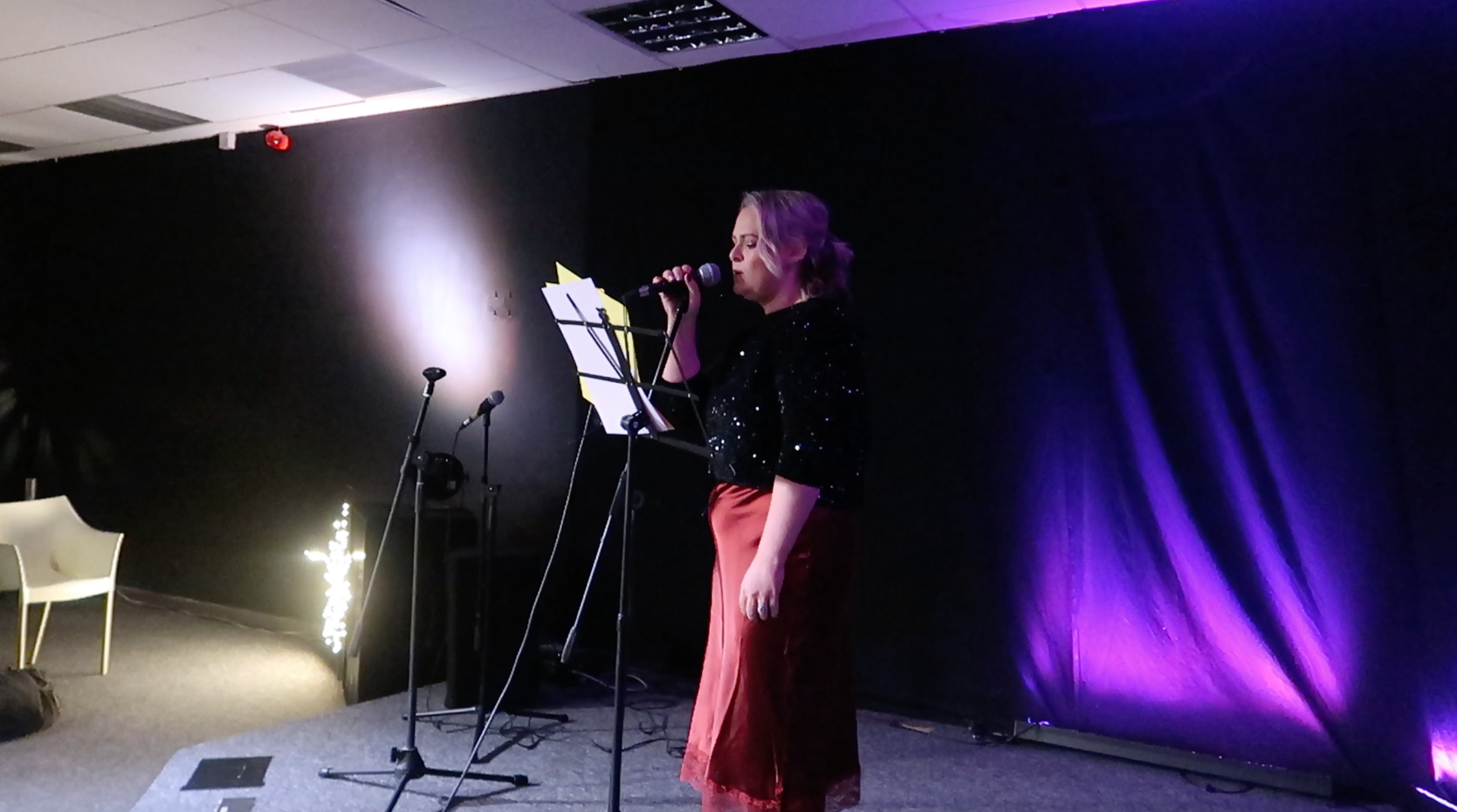 A woman singing on a stage with purple lighting, holding a mic, wearing a black top with sequins and a red skirt.