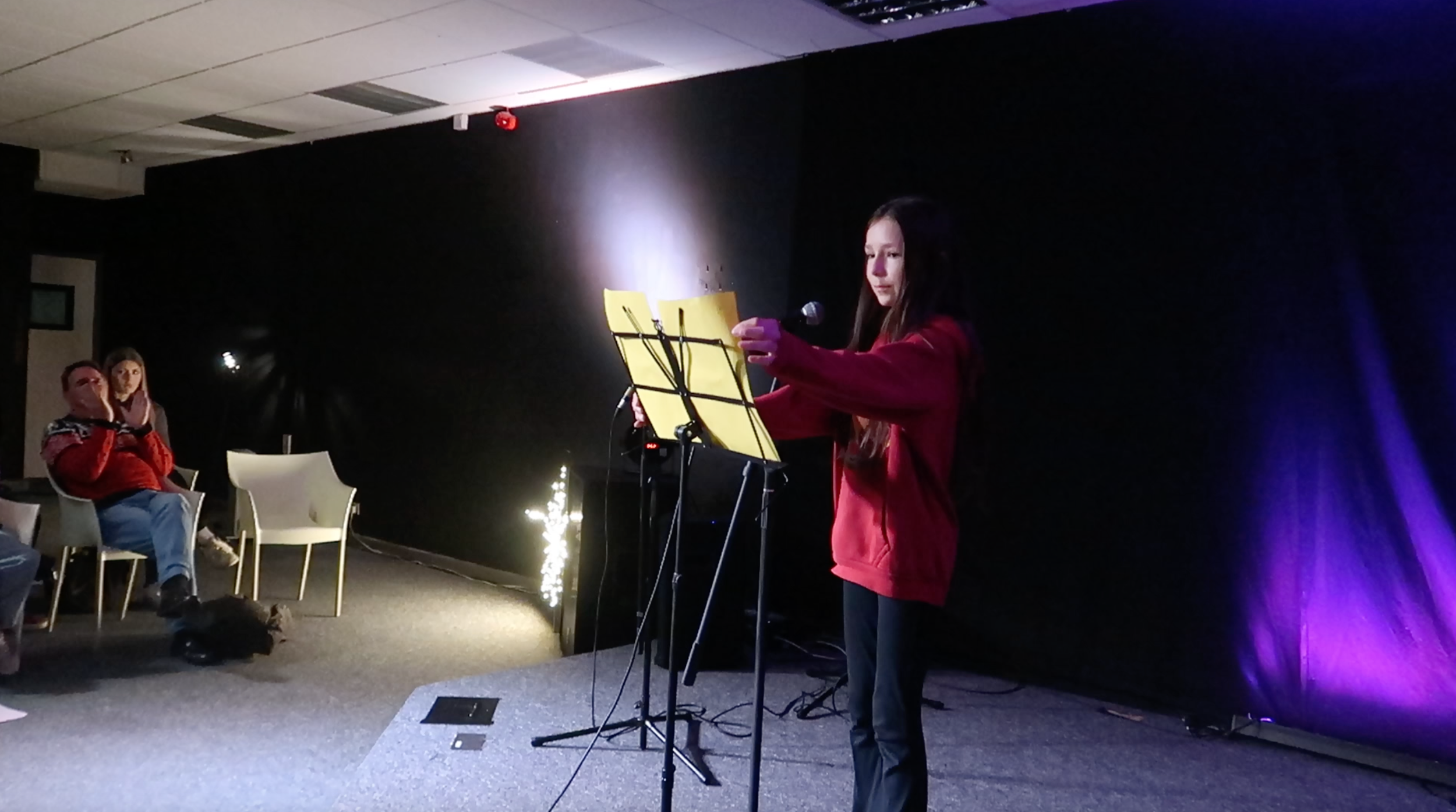 A young woman performing on stage with a musical stand, microphone, and yellow sheet music in a dark room with purple lighting, audience members seated nearby.
