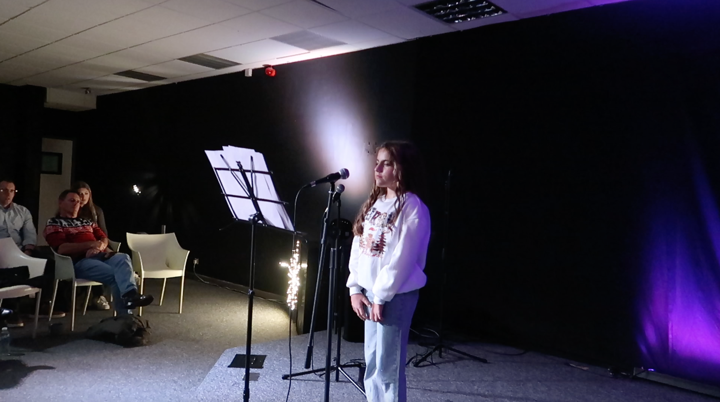 A young woman stands on stage in front of microphones, reading or singing while a small audience watches in a dimly lit room decorated with sparklers.