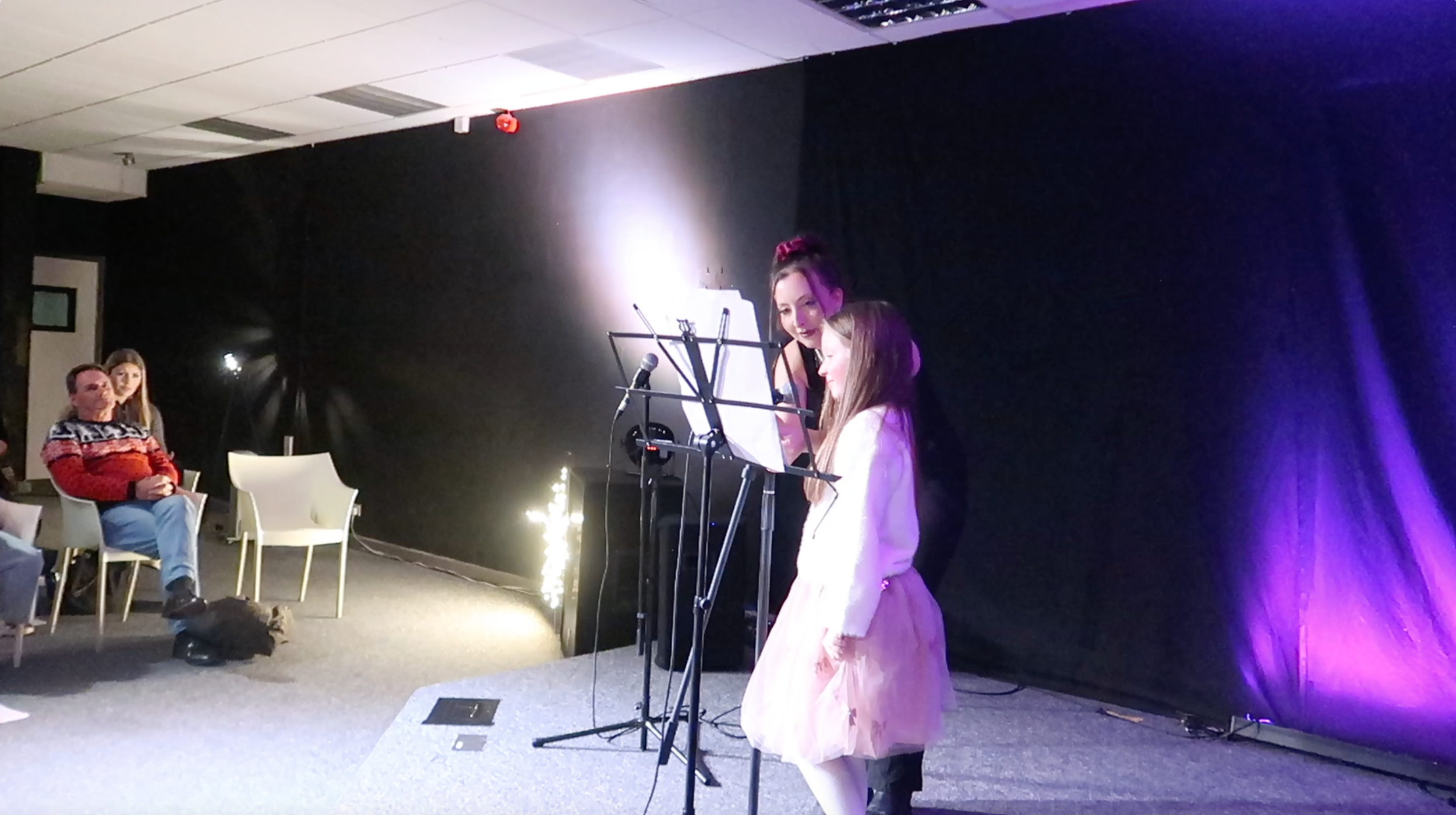 Two young girls are singing or speaking into microphones on stage while an audience watches. A woman stands behind them, possibly assisting. The stage has purple lighting and a black backdrop, and some audience members are seated nearby.