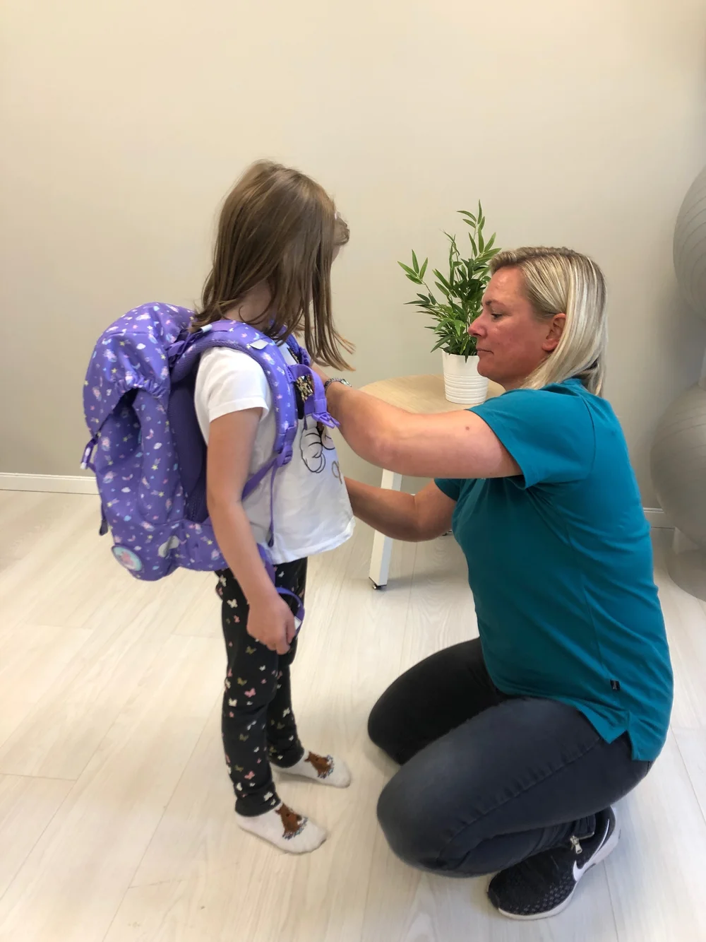 En voksen justerer stroppene på en lillamønstret ryggsekk som bæres av en ung person i et sterkt opplyst innendørsrom.