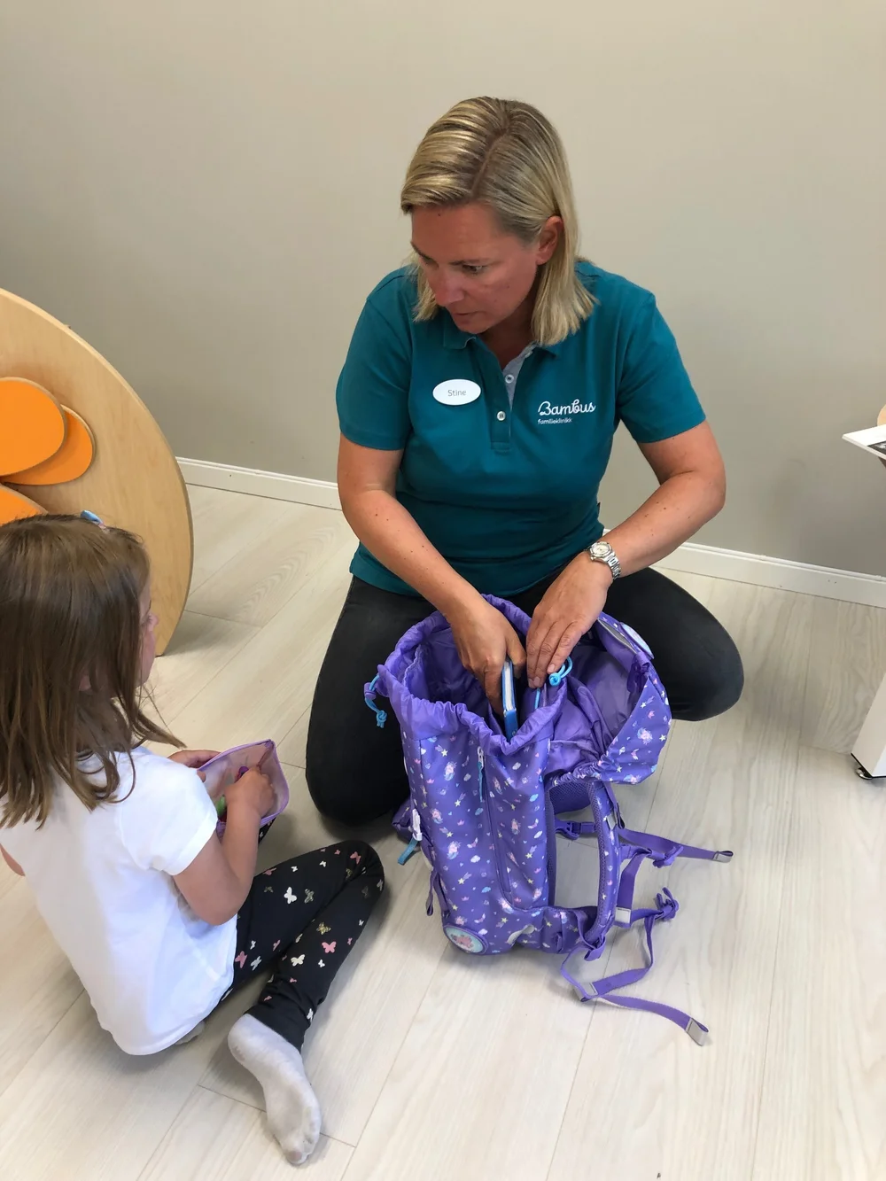 En person i en blågrønn skjorte kneler på et lyst gulv og hjelper et barn med å plassere en gjenstand i en lillamønstret ryggsekk.