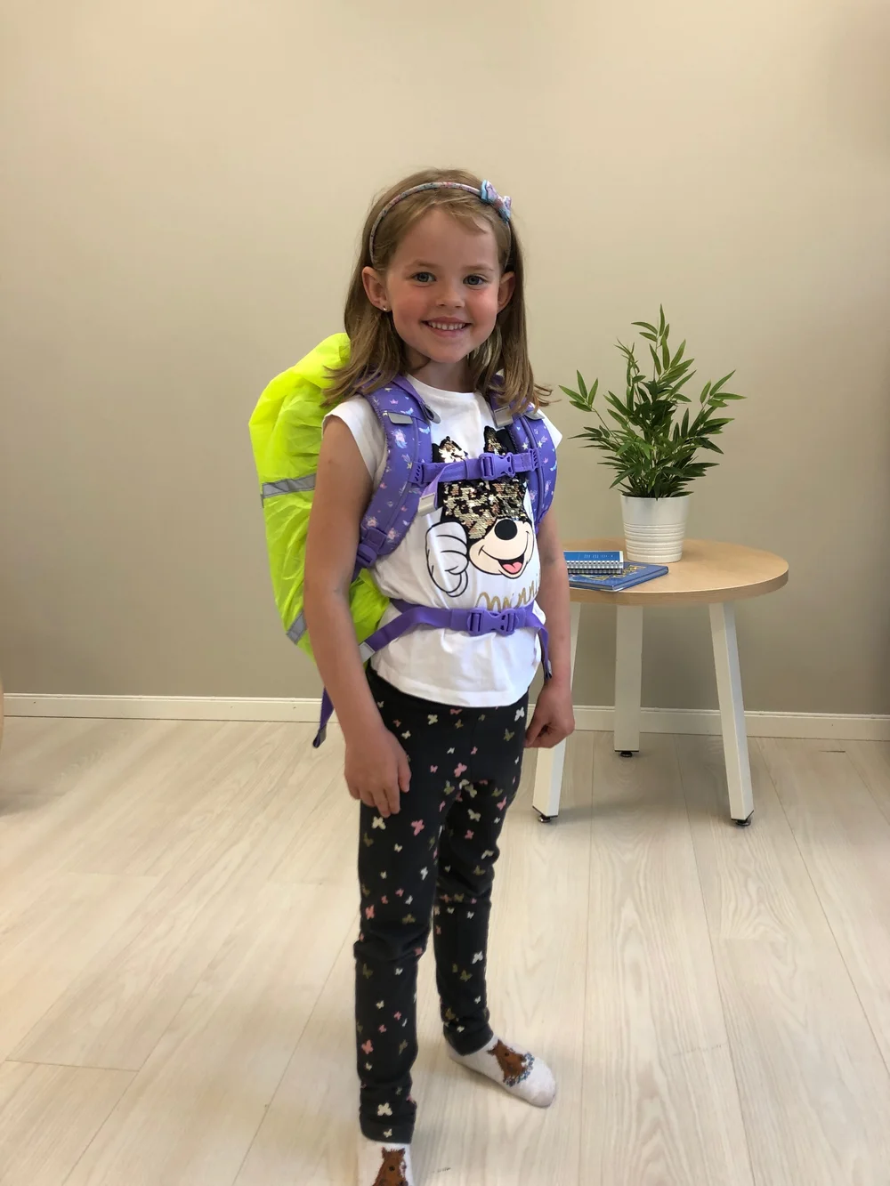 Et smilende barn har på seg en knallgul ryggsekk og en hvit t-skjorte med trykk innendørs ved et lite bord med en potteplante.