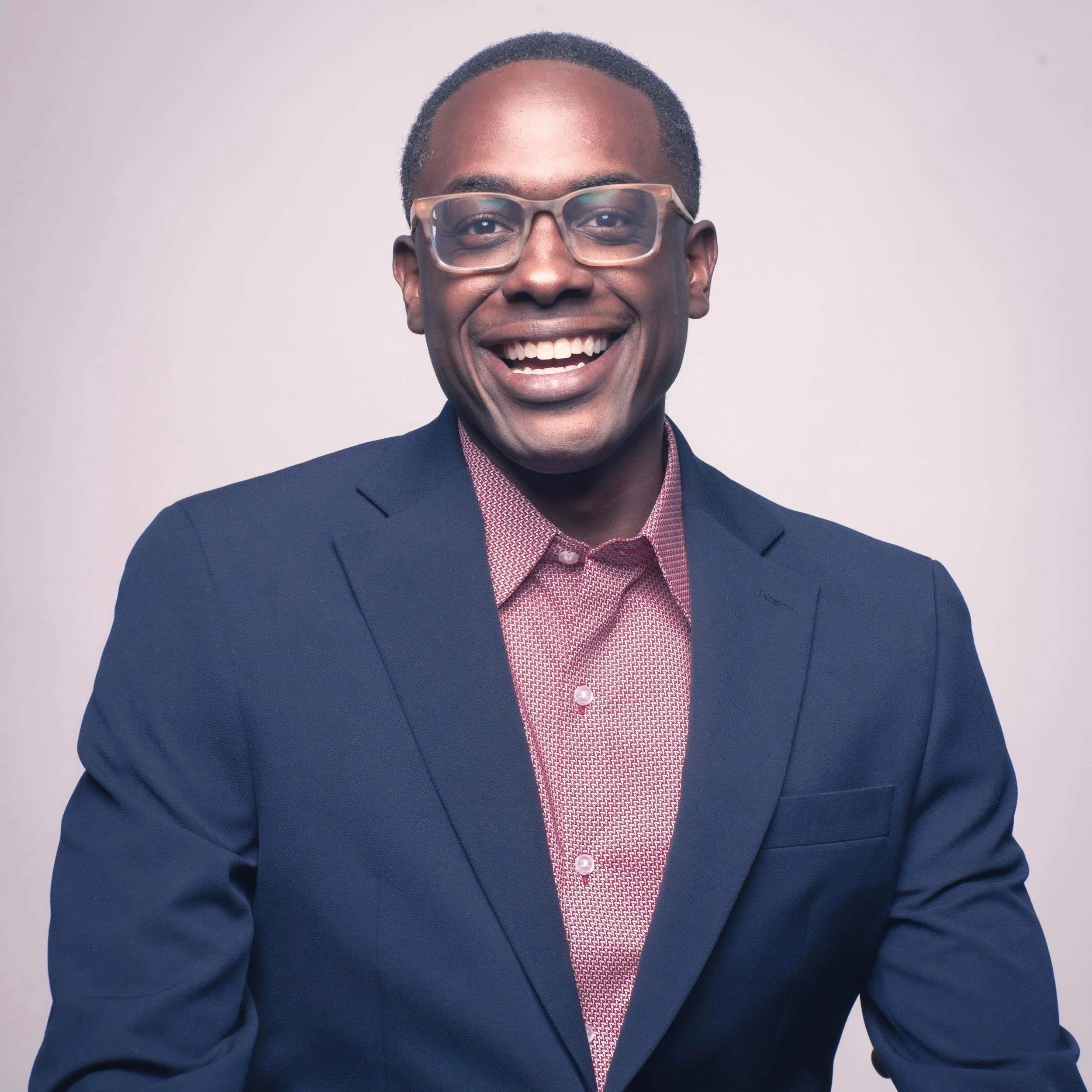 Portrait of a smiling man wearing glasses, a navy blazer, and a red patterned shirt against a plain background.
