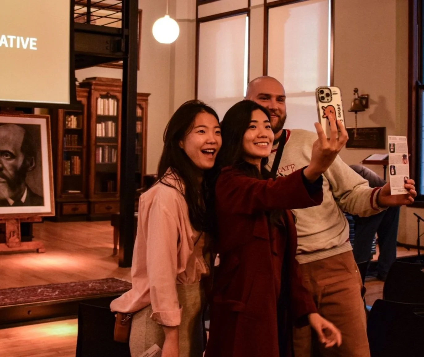 Three people taking a selfie in a room with bookshelves and a framed portrait in the background.