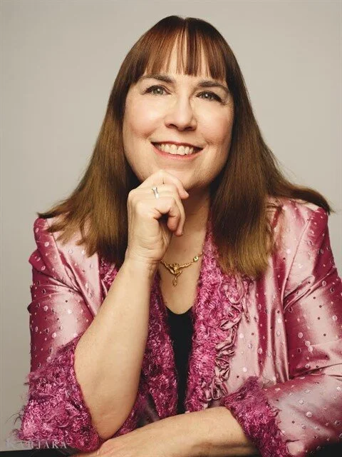 A woman with shoulder-length brown hair and bangs, smiling, wearing a pink satin blazer with ruffle details and a black top, posed with her chin resting on her hand against a plain neutral background.