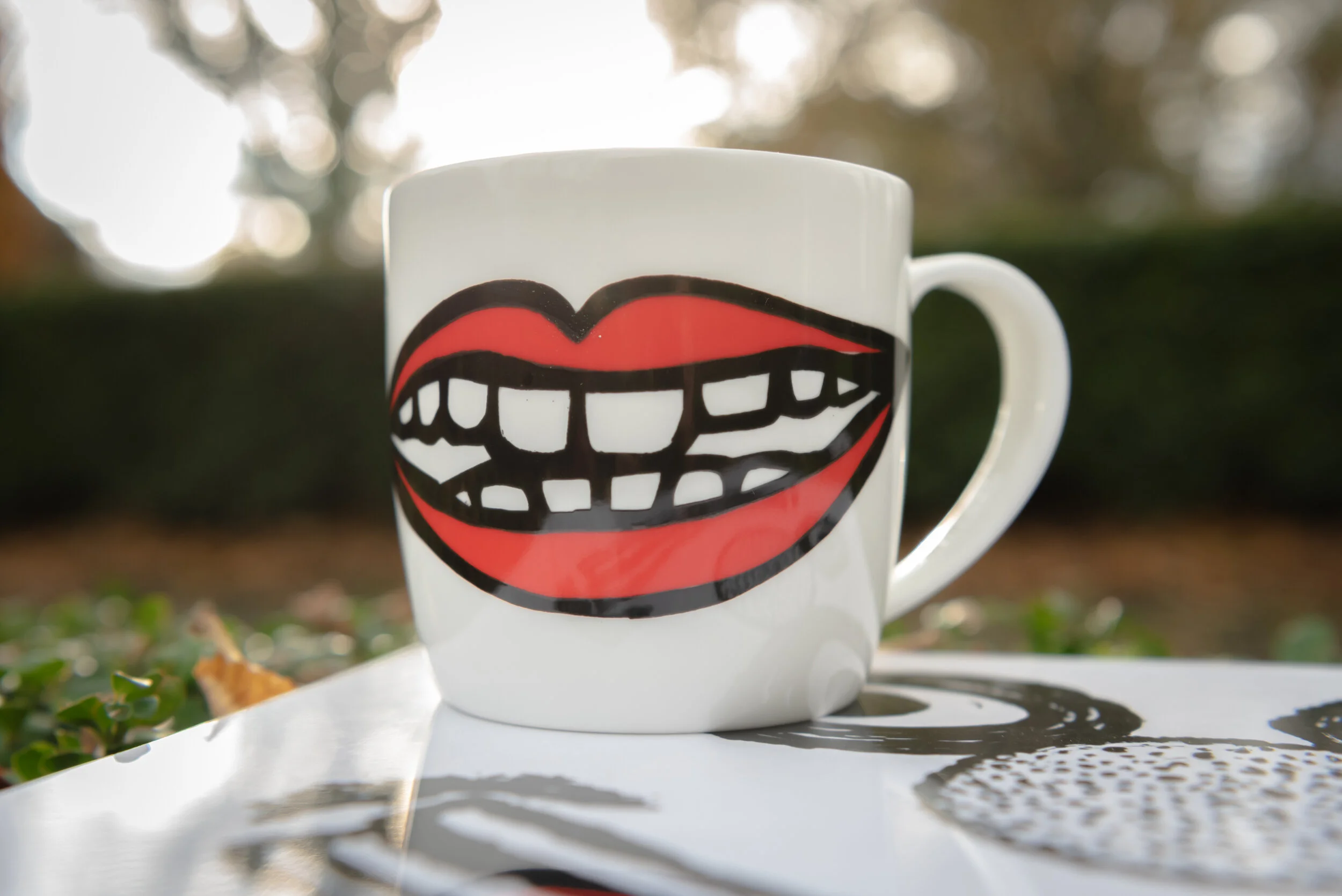 White coffee mug with a cartoon of smiling red lips and teeth on a table outdoors
