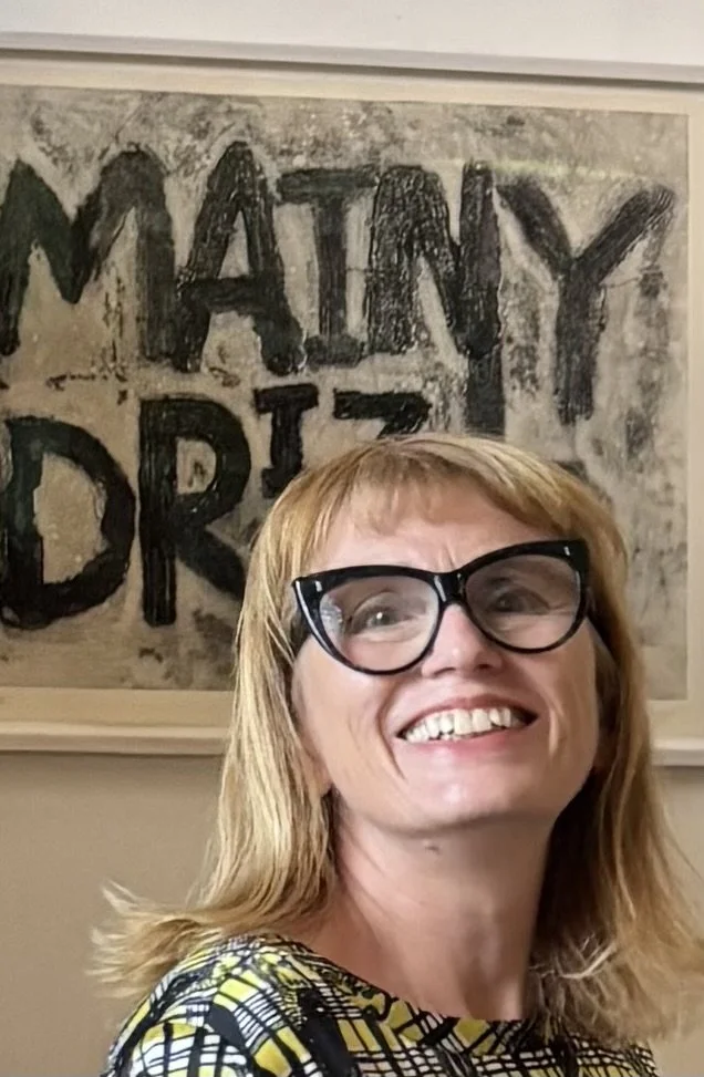 Smiling woman wearing black glasses in front of an abstract artwork reading “MATIN”