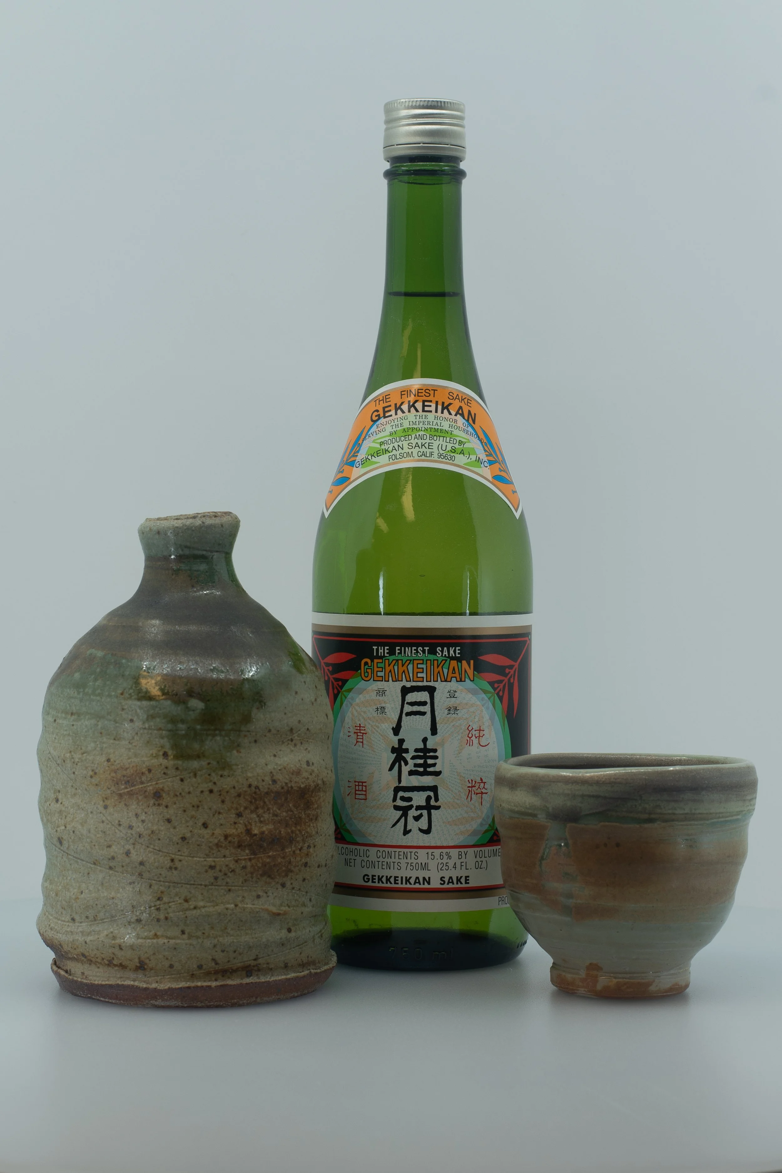 A green bottle of Gekkeikan sake, flanked by two ceramic sake cups with a rustic, glazed finish, against a plain, light background.
