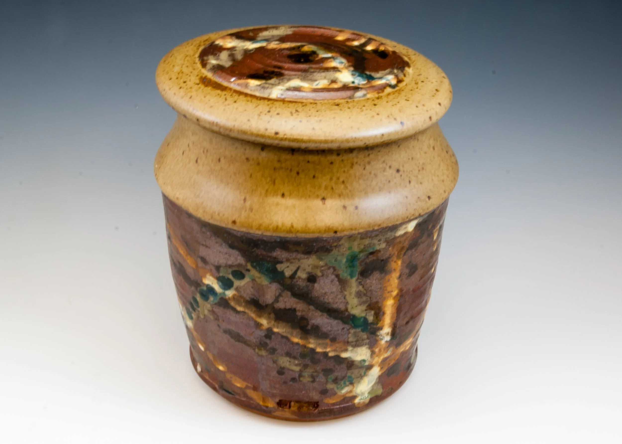 A small, rounded ceramic vase with a textured, mottled glaze in shades of brown, red, green, and gold, featuring an abstract, swirling pattern on the top lid.