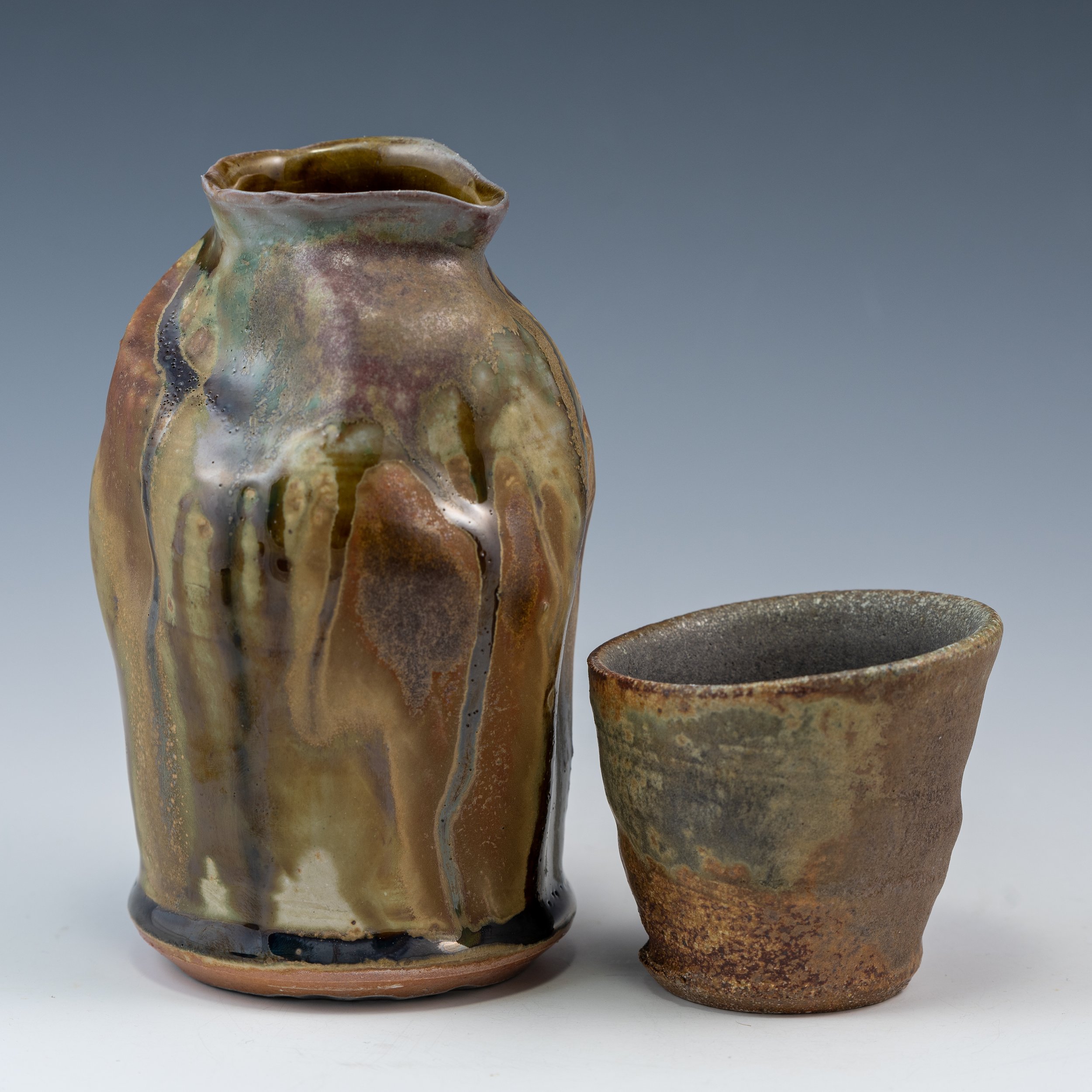 Two handmade ceramic vessels with earthy tones and textured glazes, one taller with a ruffled opening and the other shorter with a rounded opening, set against a plain background.