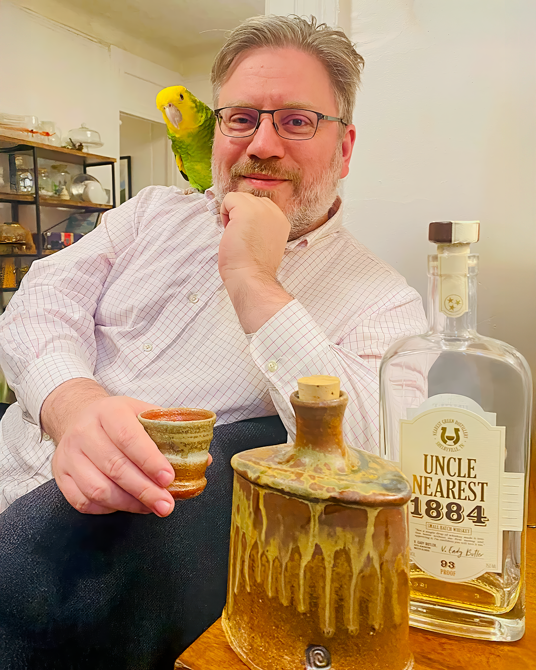 A man with glasses and a beard sitting at a table, with a green parrot on his shoulder, holding a small ceramic cup near a large bottle of Uncle Nearest whiskey, with decorative ceramic items on the table and a background of shelves with glassware.