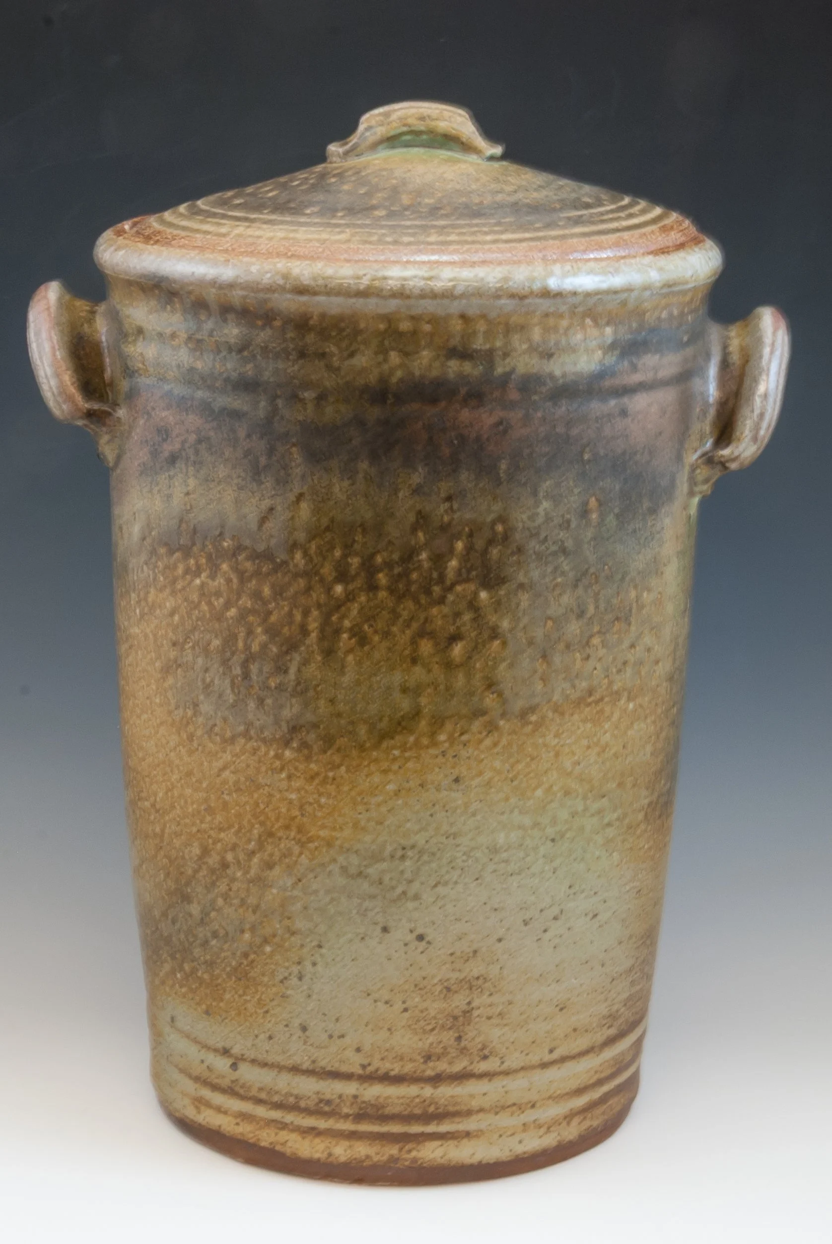 Ceramic vase with a lid, featuring a textured and gradient glaze in earthy tones of brown, beige, and green, with a rounded shape and small handles on the sides.