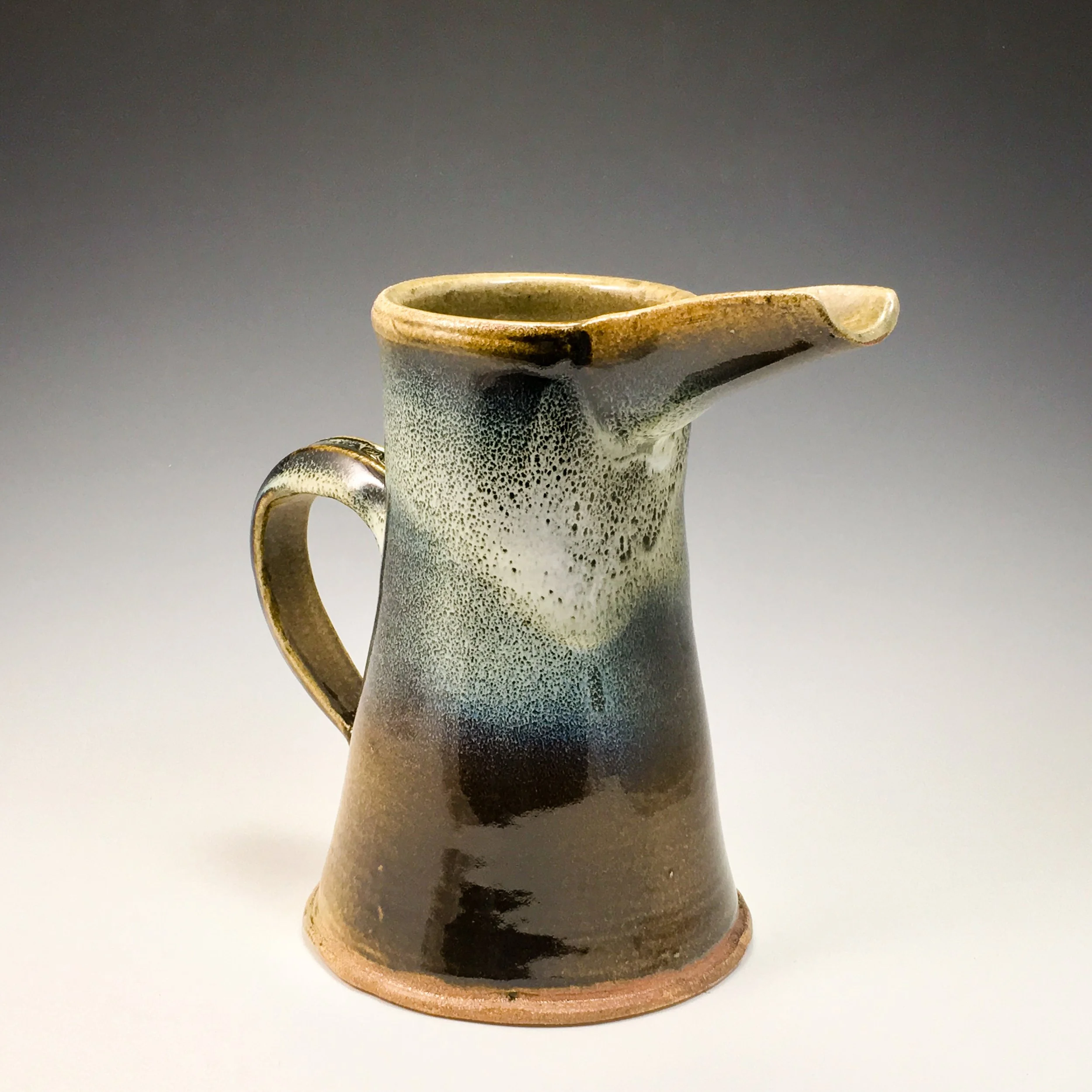 A ceramic pitcher with a speckled gray and brown glaze, featuring a wide spout and a handle, placed on a white surface.