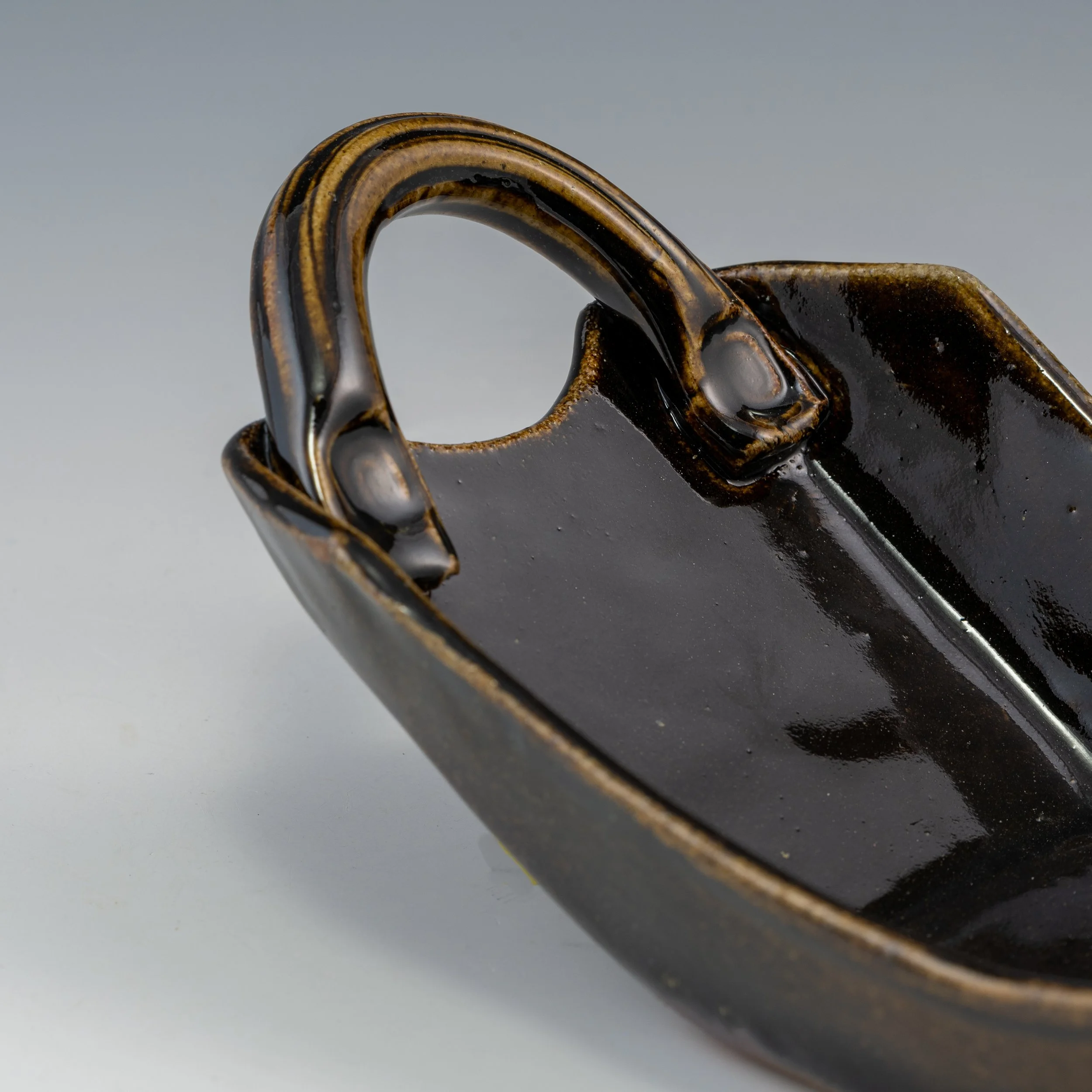 Close-up of a ceramic mug with a glossy dark glaze and a decorative brown and black band around the handle, against a gray background.