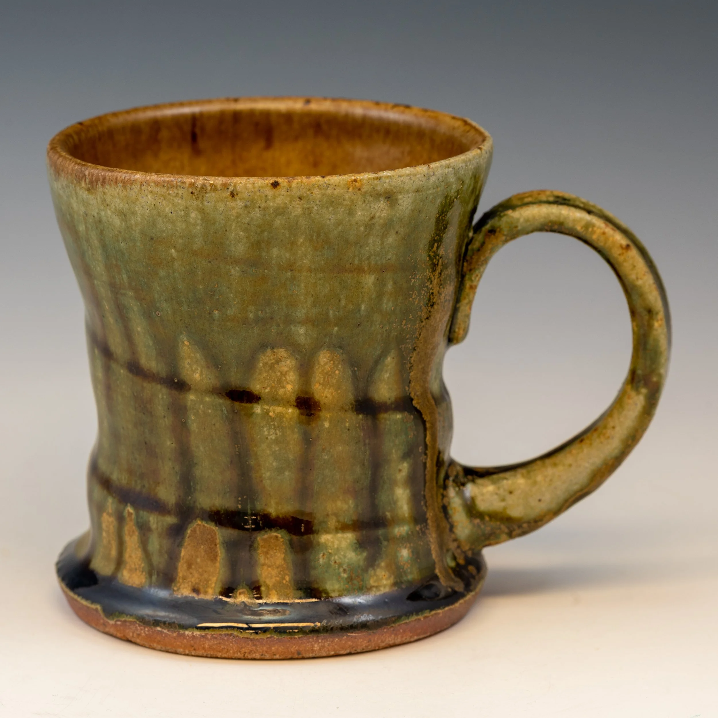 A close-up view of a small, ancient ceramic cup with a rounded handle. The cup has an earthy, greenish-brown glaze with a distressed, weathered surface, revealing signs of age and use.