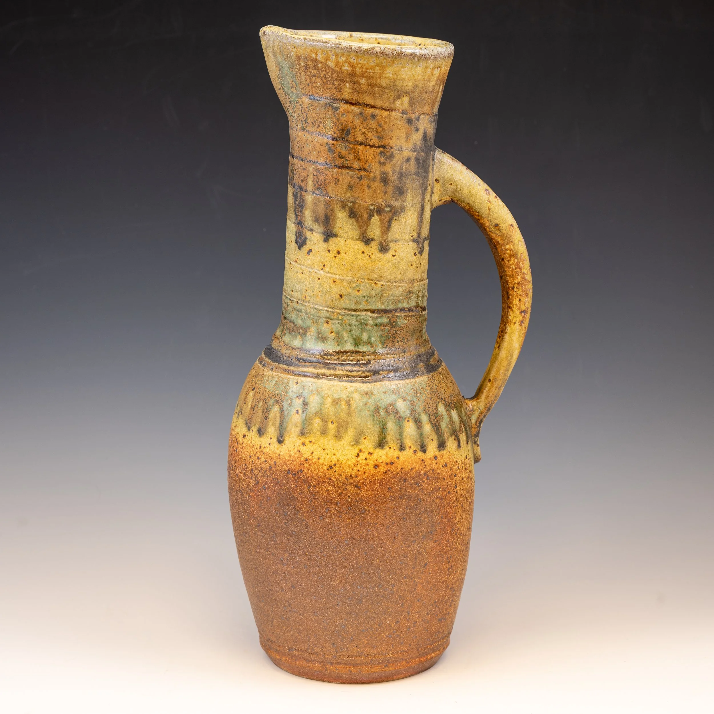 A ceramic vase with a tall, narrow, cylindrical body, a rounded lip, and a small handle on the side. The vase has a textured, speckled surface with earthy tones of brown, orange, and green, and a weathered appearance.