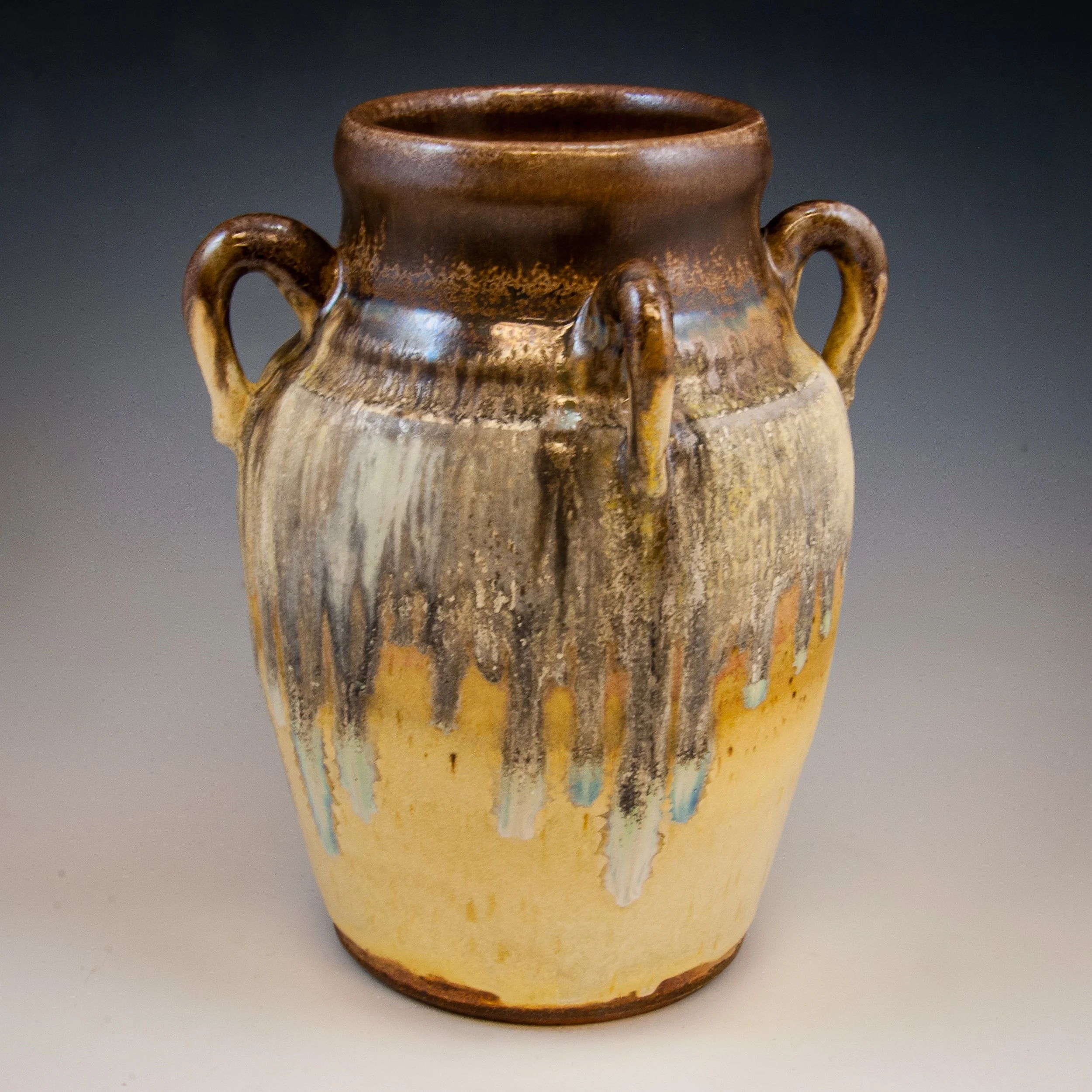 Close-up of a ceramic vase with a glossy brown top and earthy tones, featuring four small handles and a textured, multicolored surface, set against a dark background.