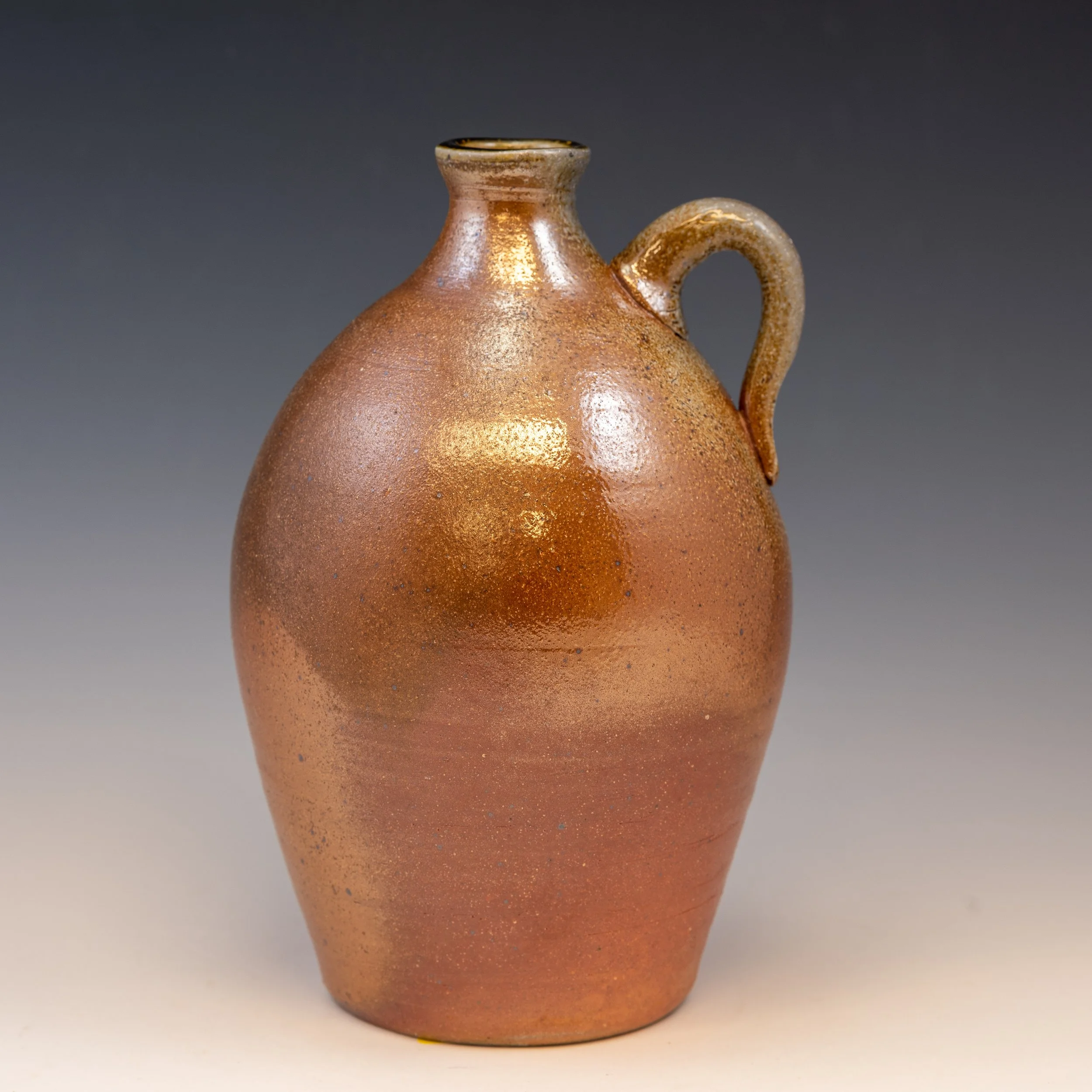 A small ceramic vessel with a round body, narrow neck, and a curved handle on the right side, featuring a reddish-brown, textured surface, displayed against a plain dark grey background.