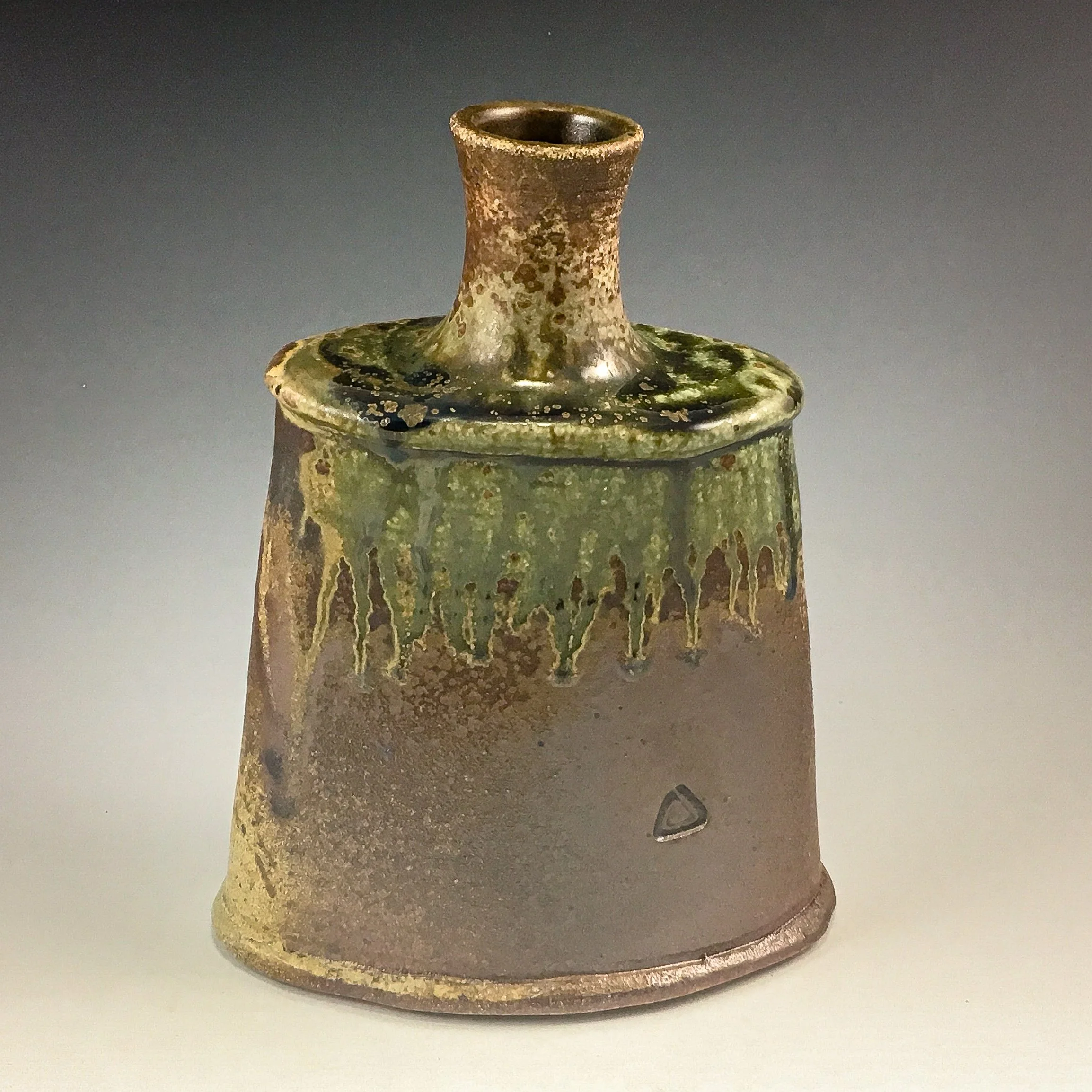 Close-up photograph of an ancient glass vessel with a narrow mouth and a wide, decorated base, featuring a mottled green and brown glaze.