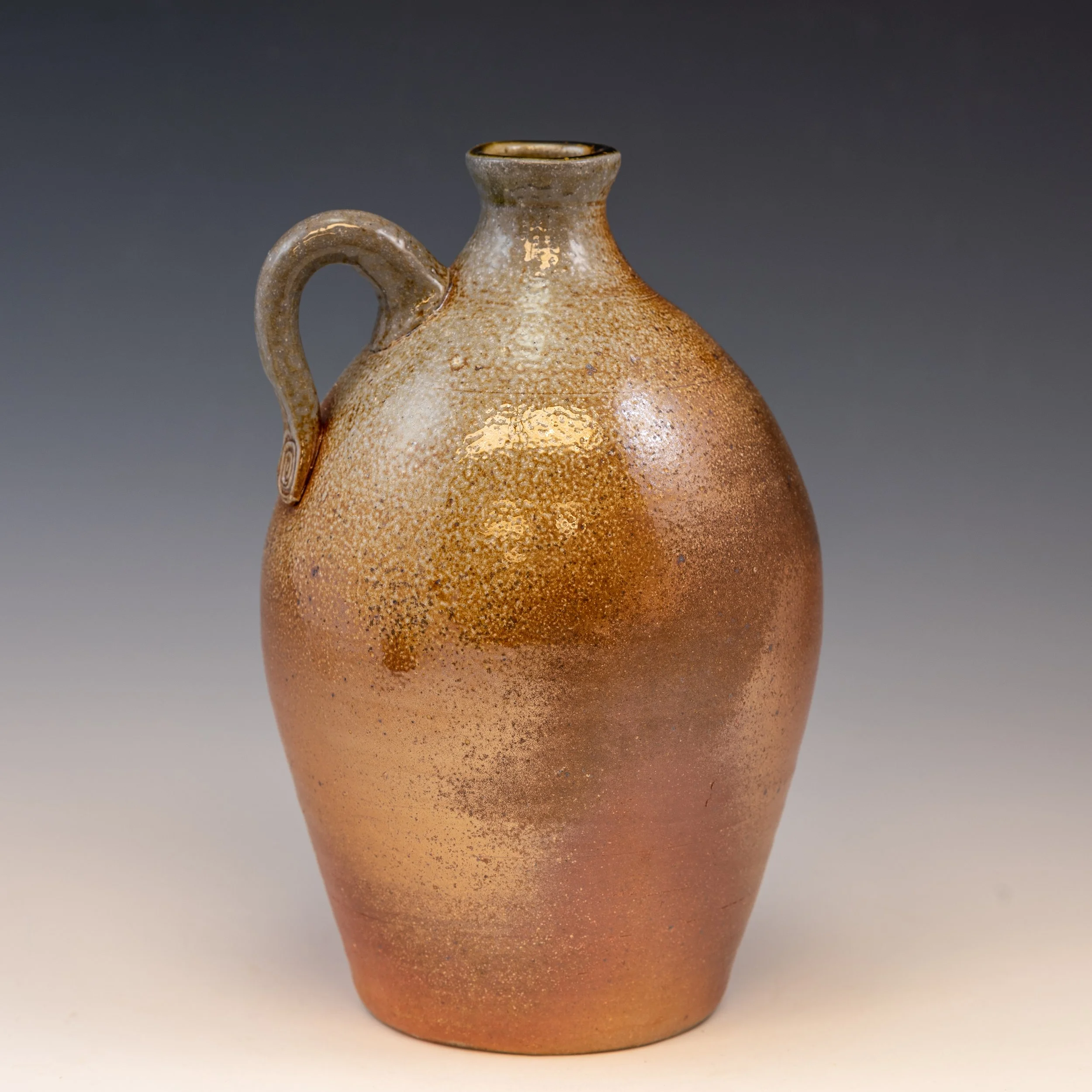 A ceramic vessel with a narrow neck, rounded body, and a small handle, featuring a textured, speckled brown glaze, set against a simple gray background.