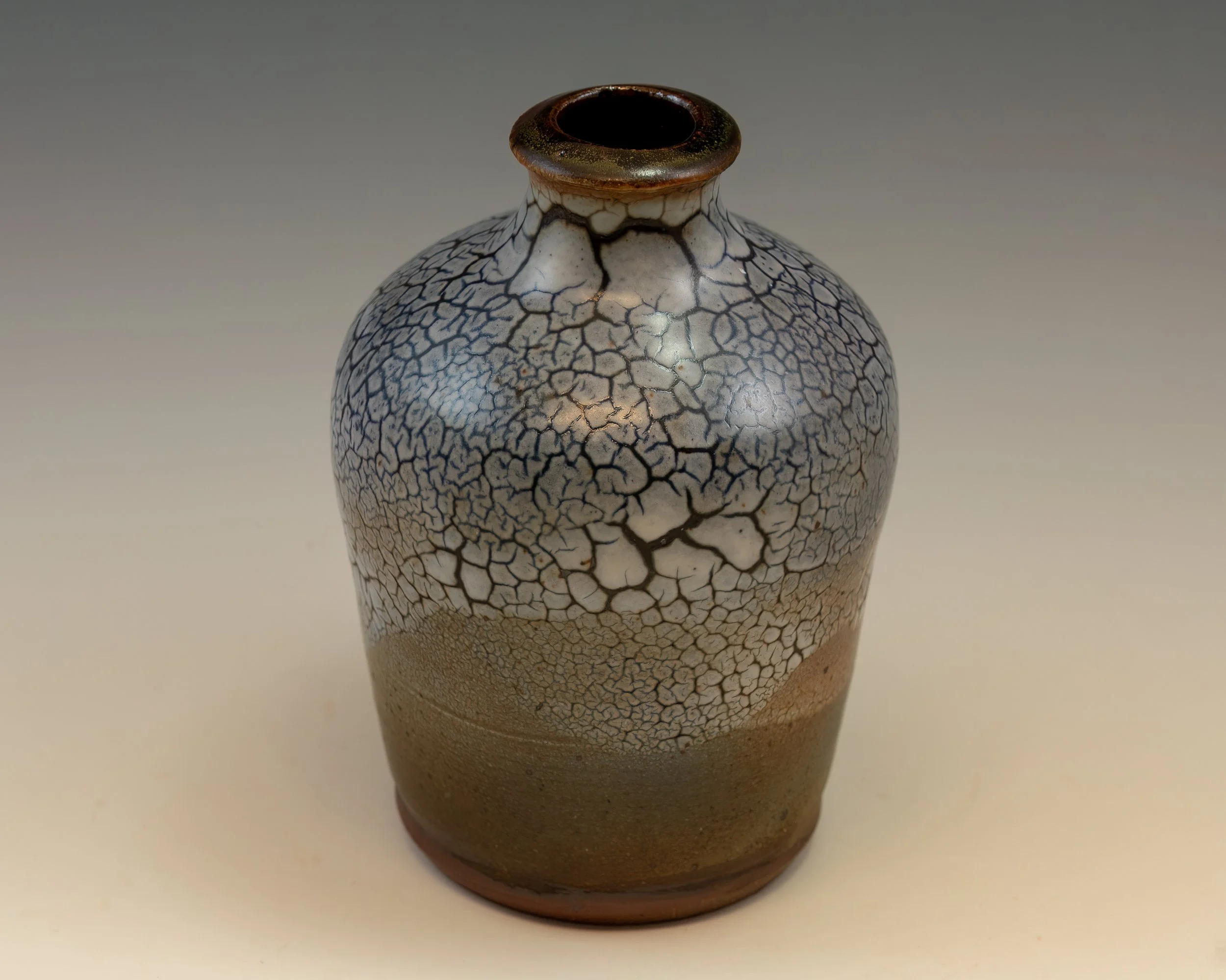 A ceramic vase with a dark, glossy rim at the top, decorated with a crackle pattern in black and white on its body, gradually transitioning to a smooth, earthy brown at the bottom.