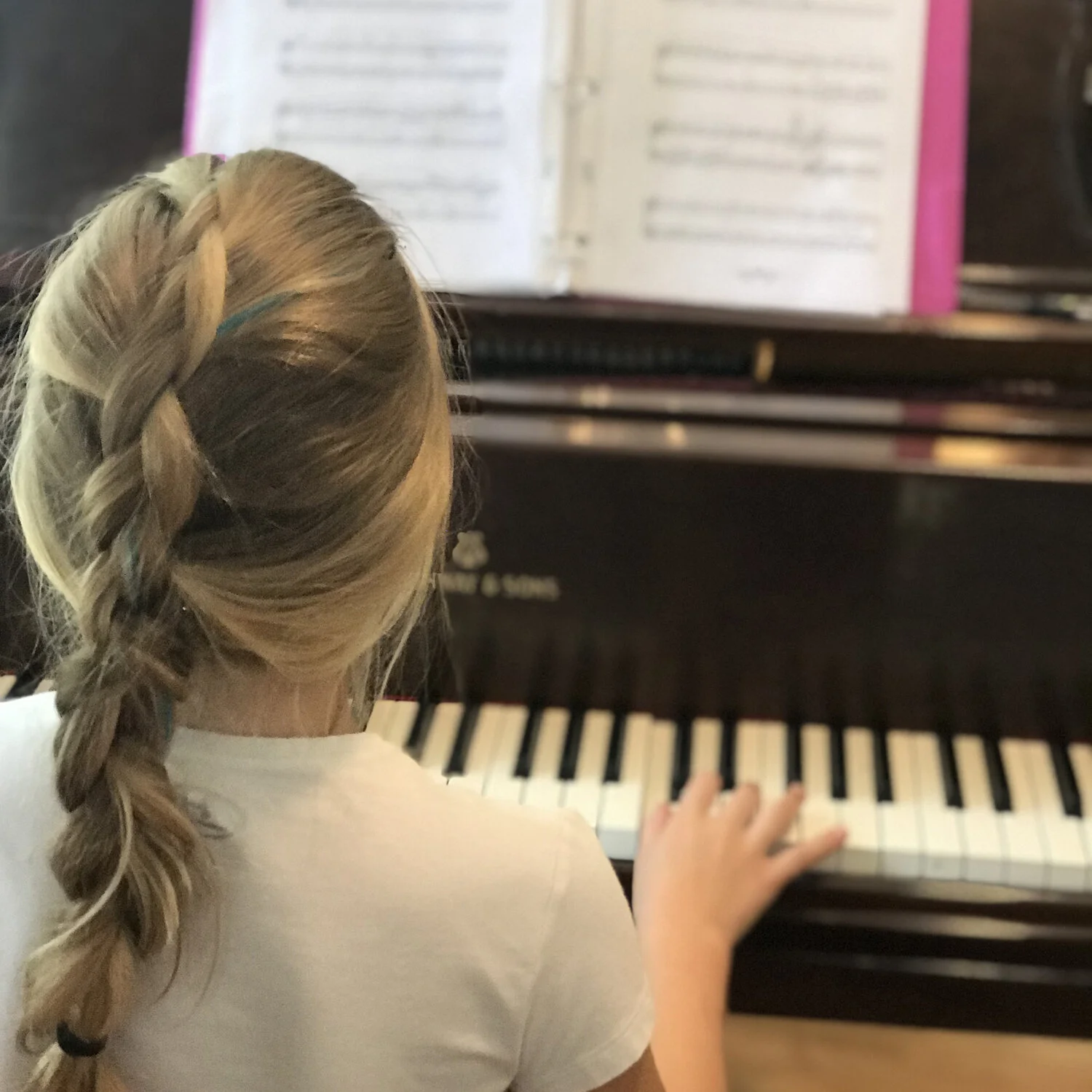 A young girl with long, braided hair playing a piano, with a music book open in front of her.