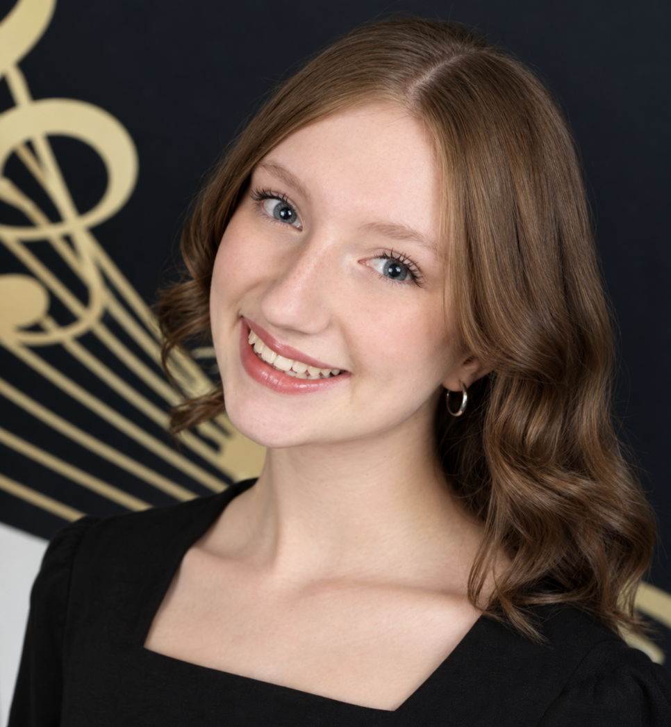 A woman with shoulder-length wavy red hair and blue eyes is smiling at the camera, wearing a black top and small hoop earrings, with a black background and a large gold musical note design.
