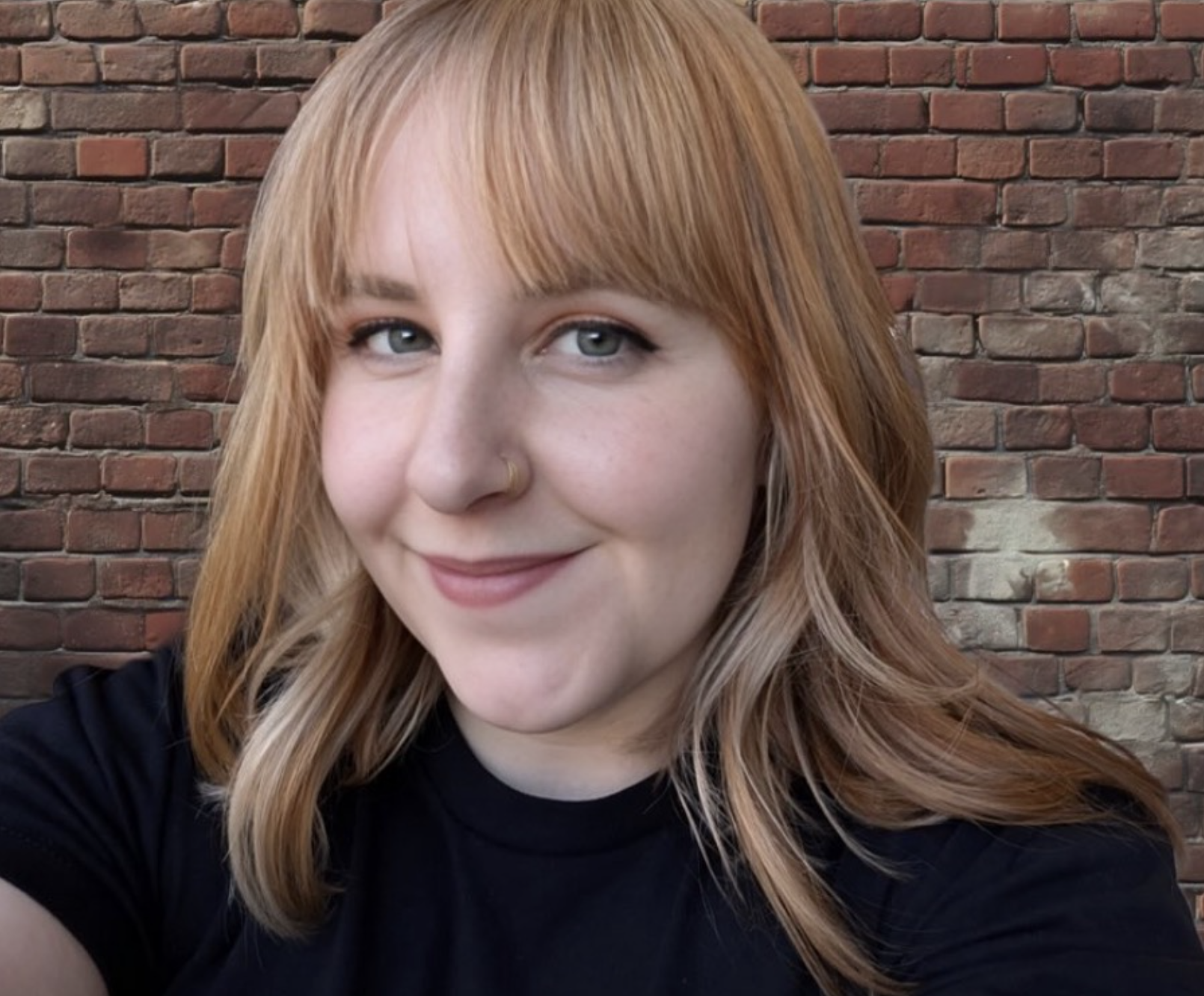 A young woman with blonde hair and blue eyes smiling at the camera, wearing a black shirt, in front of a red brick wall.