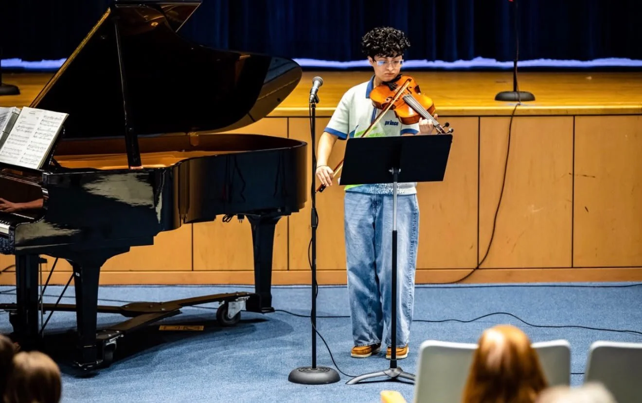 A young boy playing the violin on stage in front of an audience, with a grand piano nearby and a blue curtain backdrop.